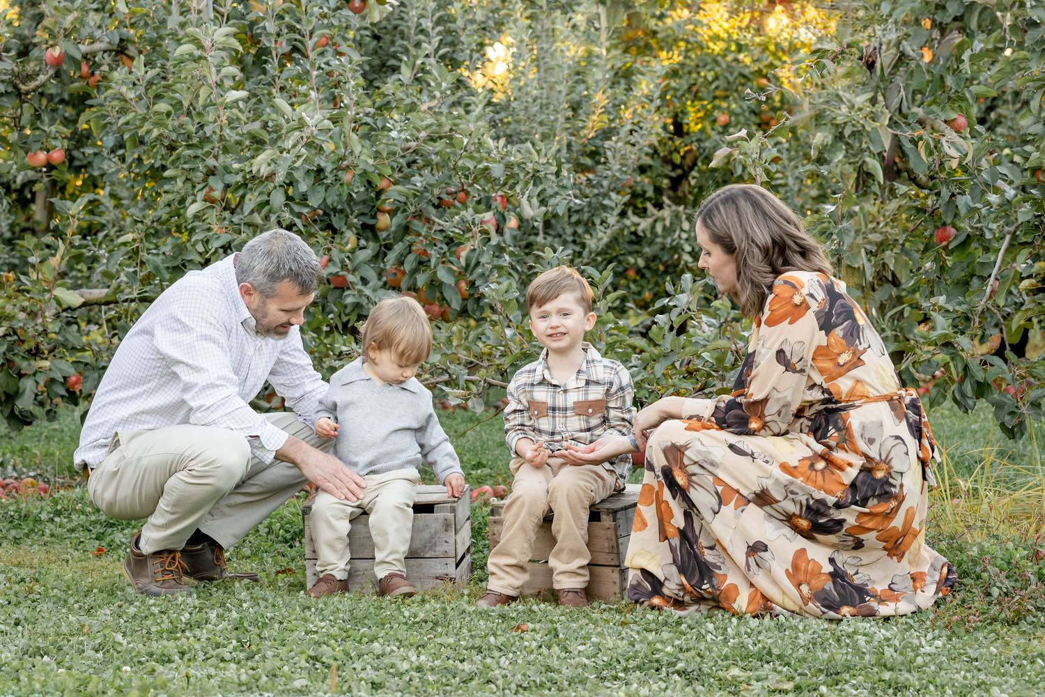 Family of four enjoying a fall day in an apple orchard, with two young boys sitting on a wooden crate.