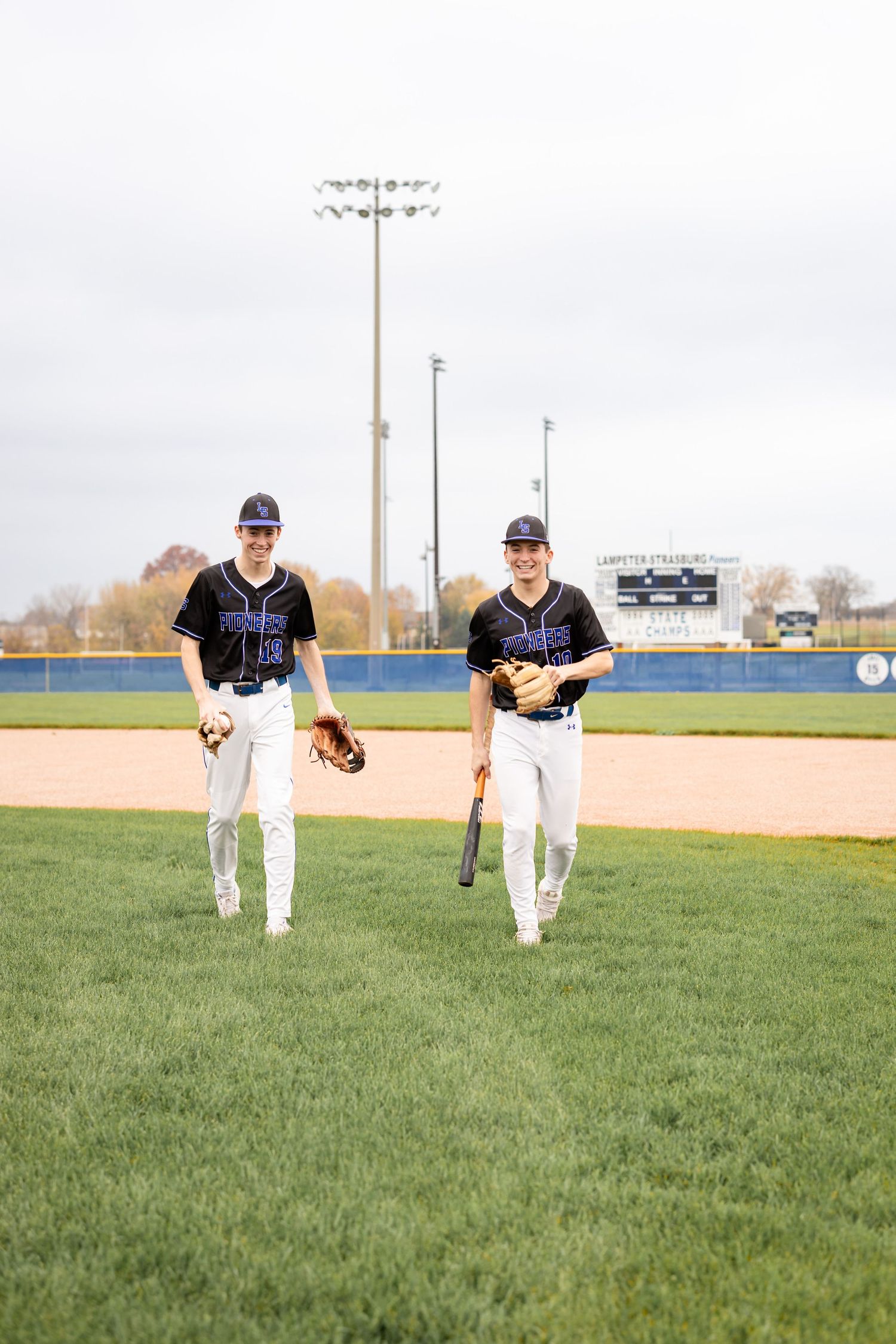 Two high school senior boys running across the baseball field in uniform during their senior photo session in Lancaster, PA