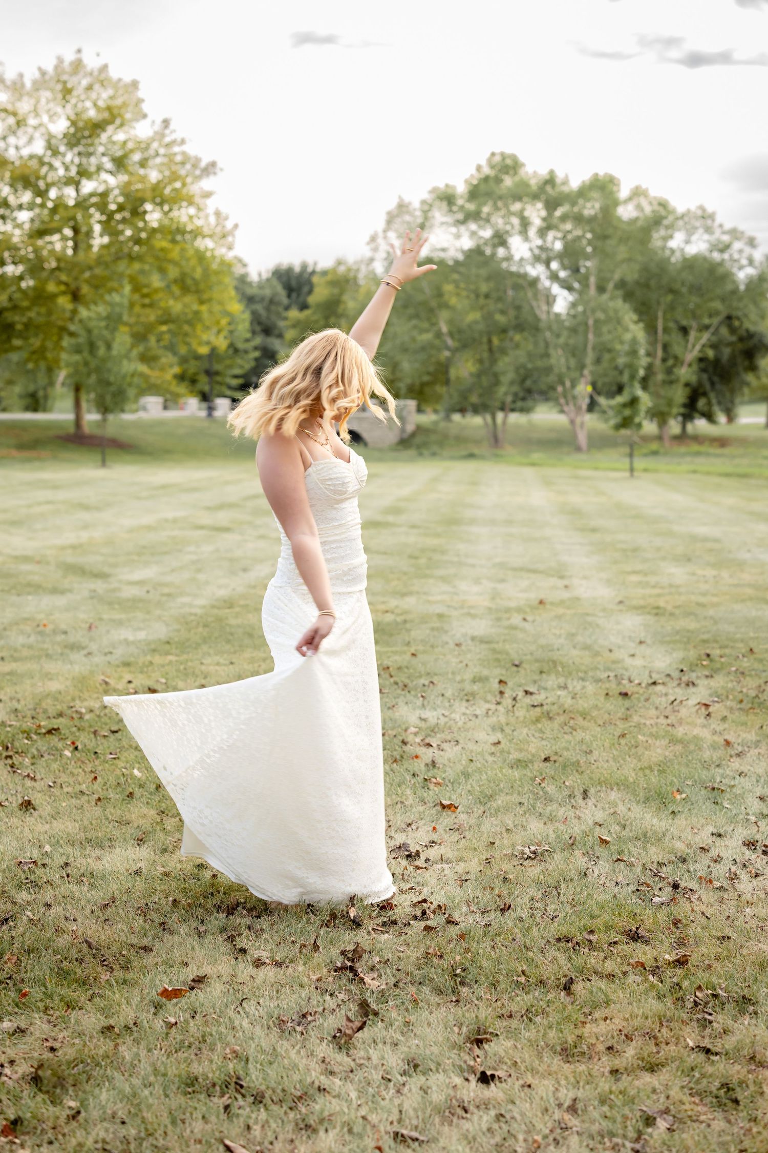 A high school senior girl in a flowing white dress twirls gracefully in a grassy field during a fall photoshoot.