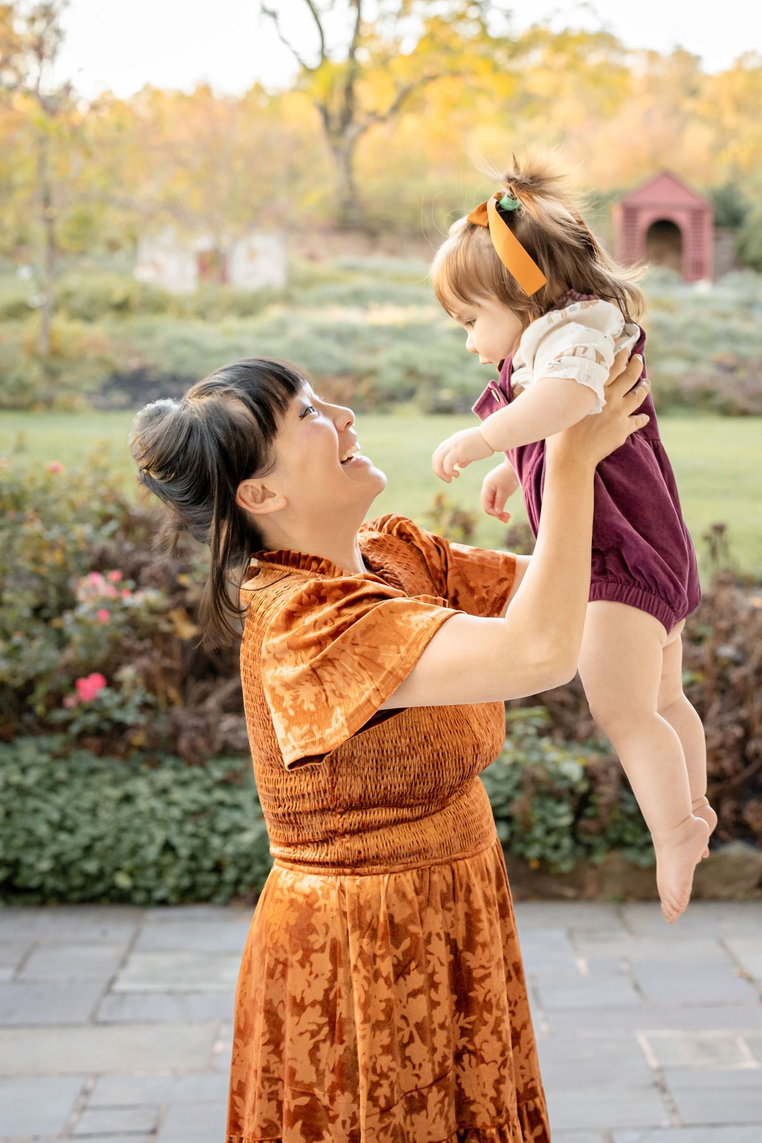 A woman in an orange velvet dress lifts a toddler girl in a burgundy romper outdoors in an autumn garden.