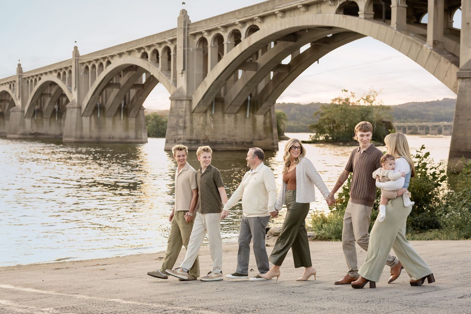 Family of seven walking along a riverfront under a stone arch bridge at golden hour.