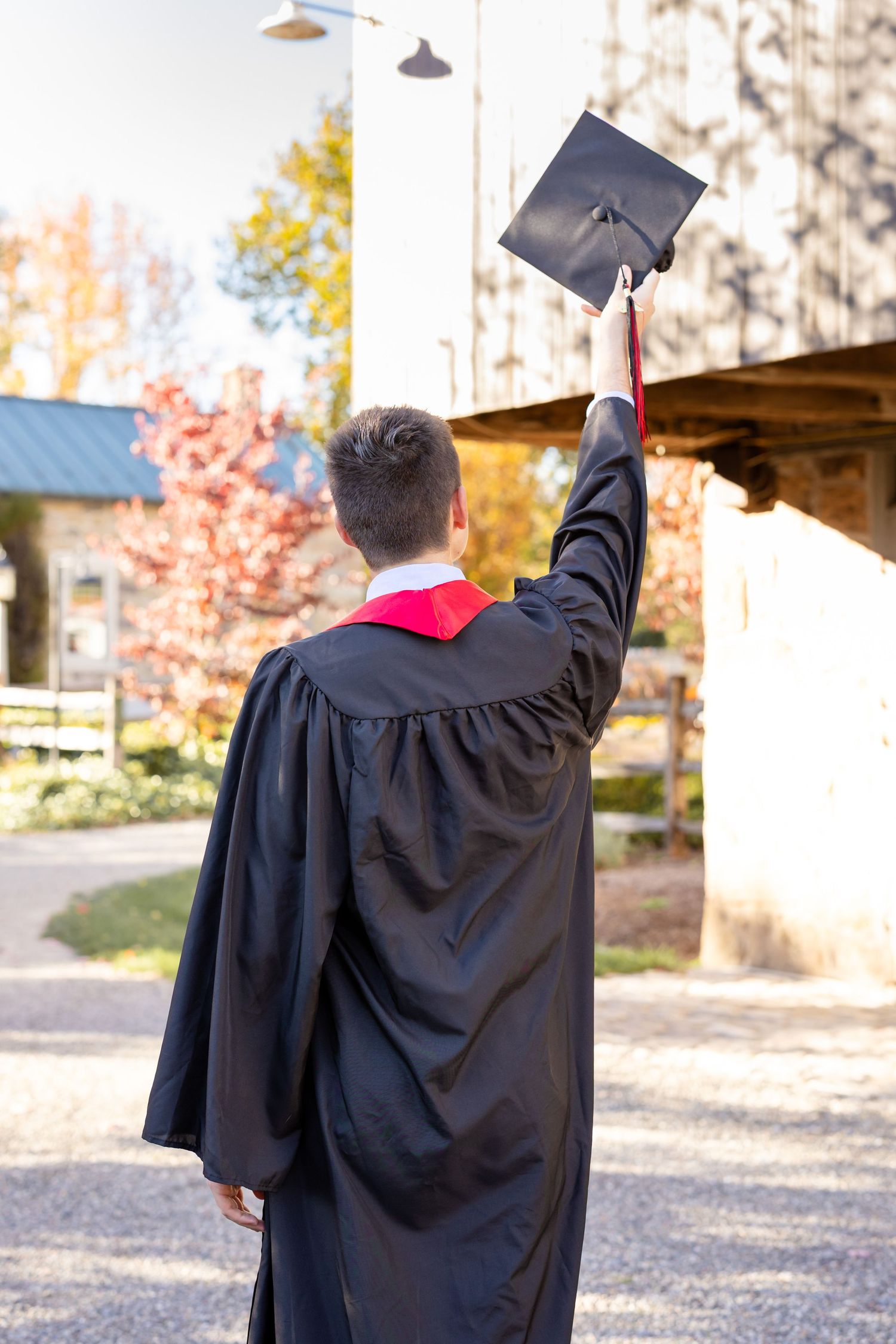 Graduate in black cap and gown raises diploma in celebration against autumn trees and building backdrop.