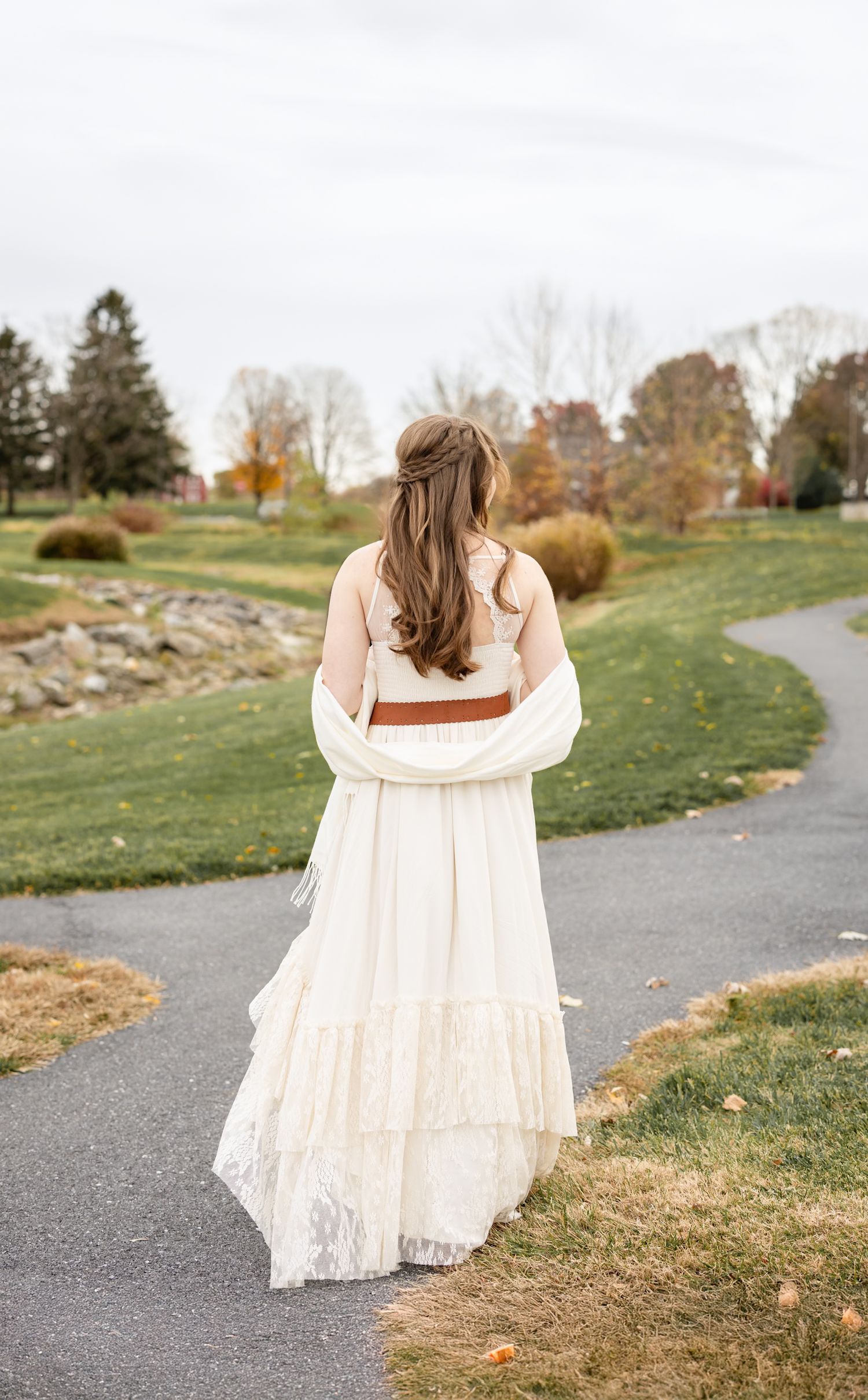 A high school graduate in a flowing white lace dress walks along a winding path surrounded by autumn grass and trees.