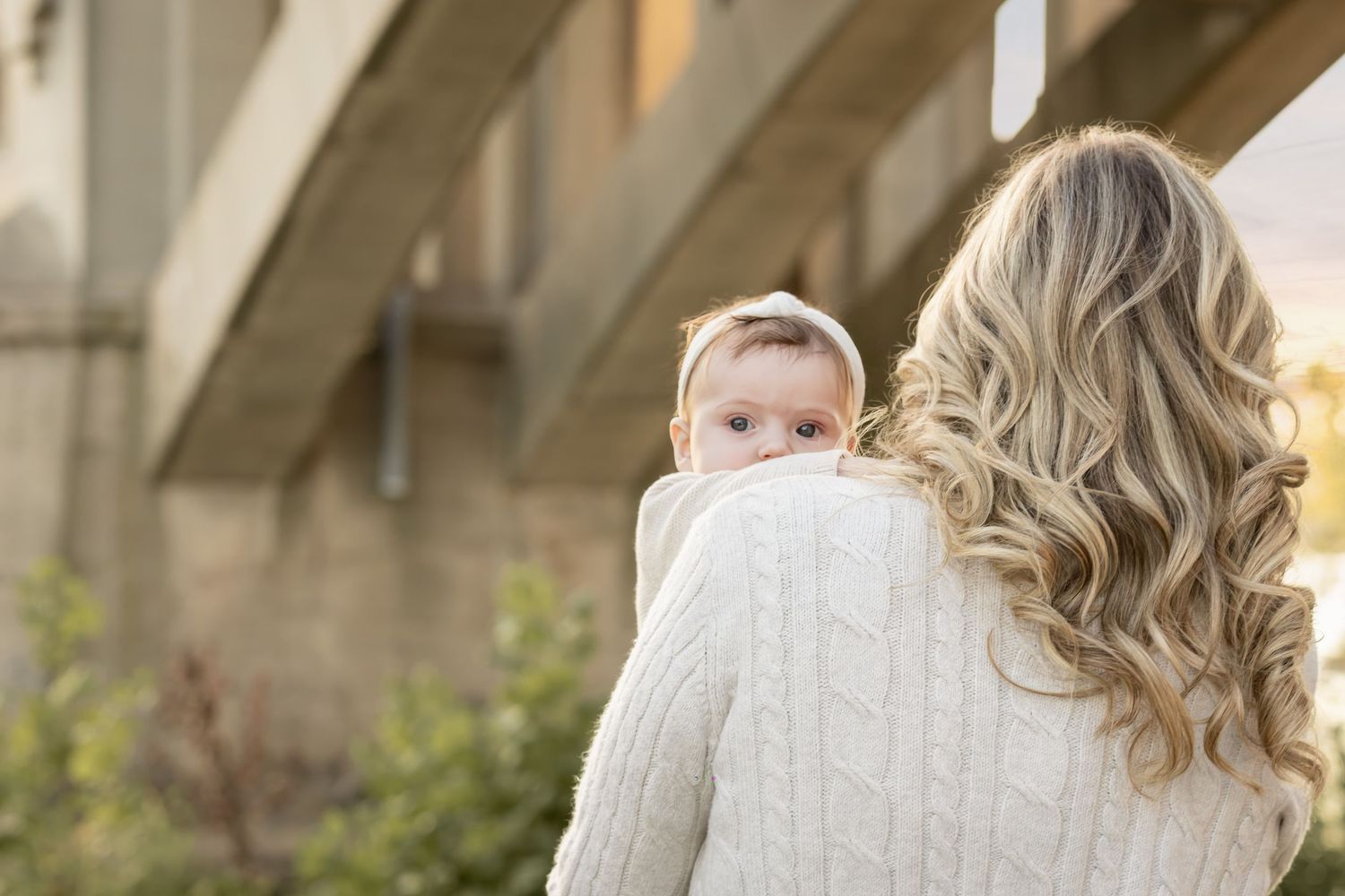 Blonde woman in cream sweater holding a baby with a bow headband near a bridge outdoors.