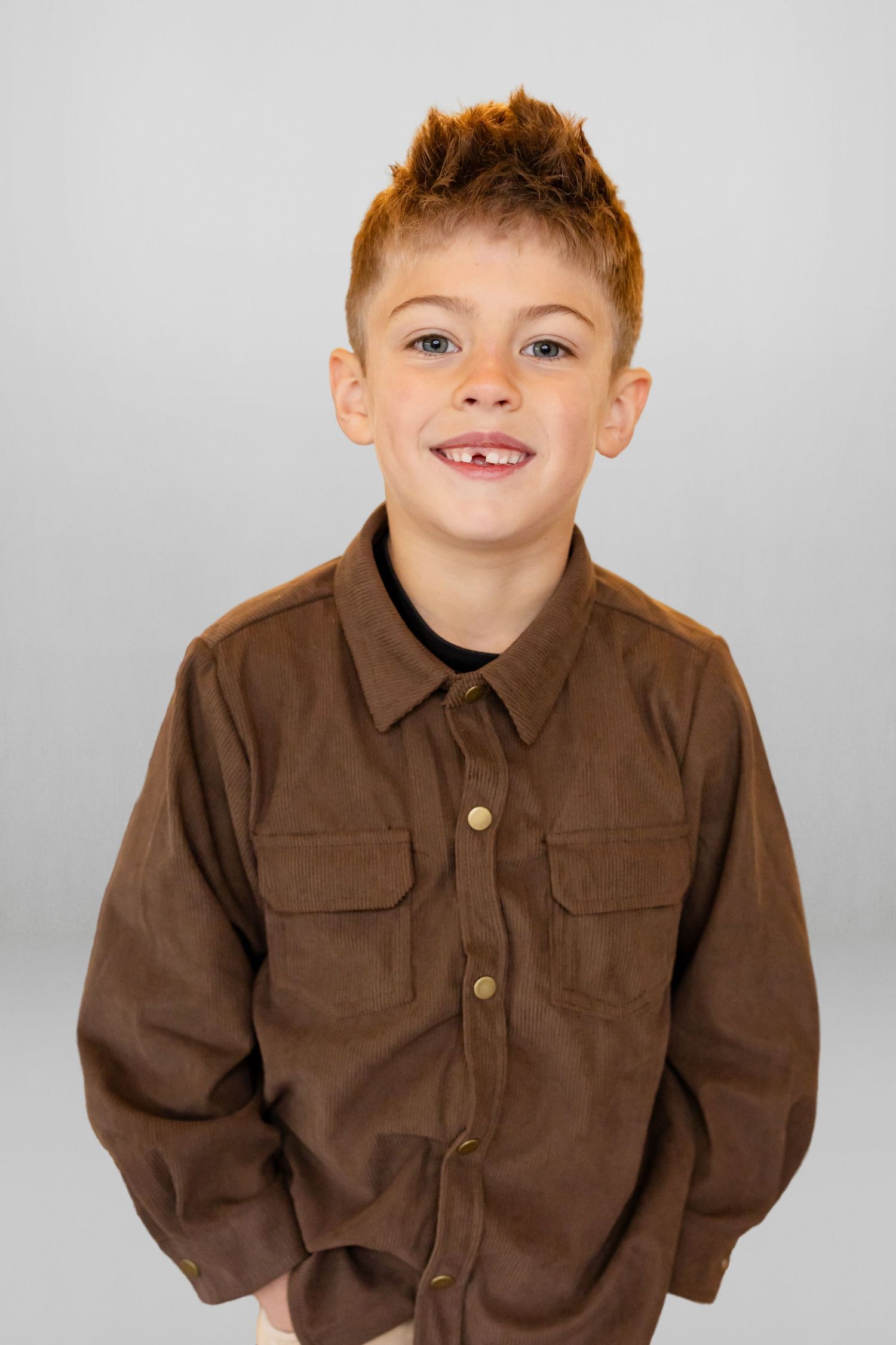 Smiling red-haired boy with a missing tooth wearing a brown button-up shirt against a gray background.