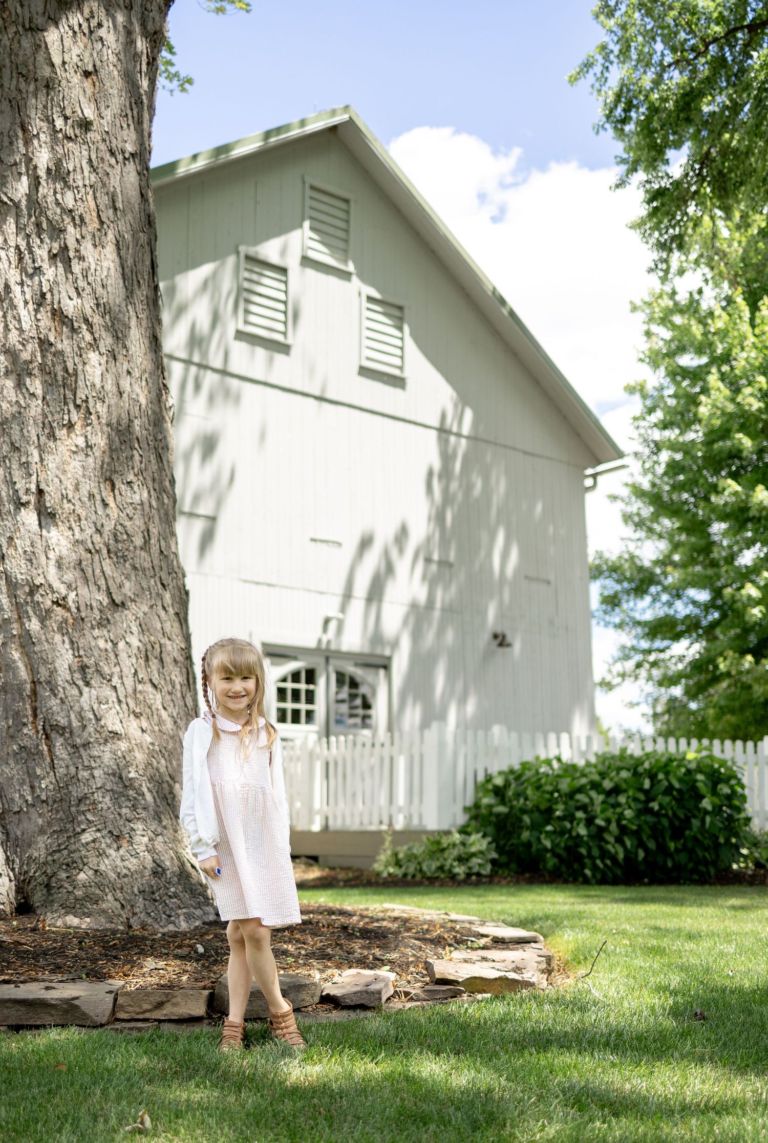 A family milestone session in Gap, PA at White Chimneys Estate. A young blonde girl in a light pink dress standing in front of a white barn.