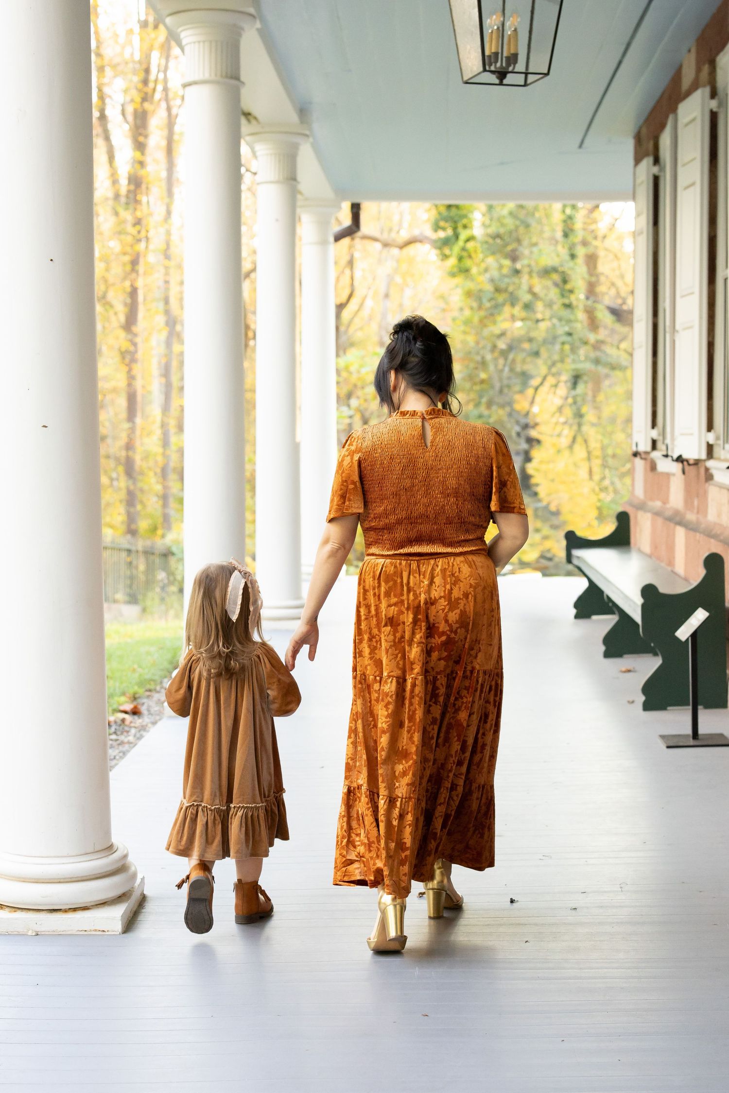 Mother and daughter in matching rust dresses walk hand-in-hand on a white porch surrounded by autumn trees.