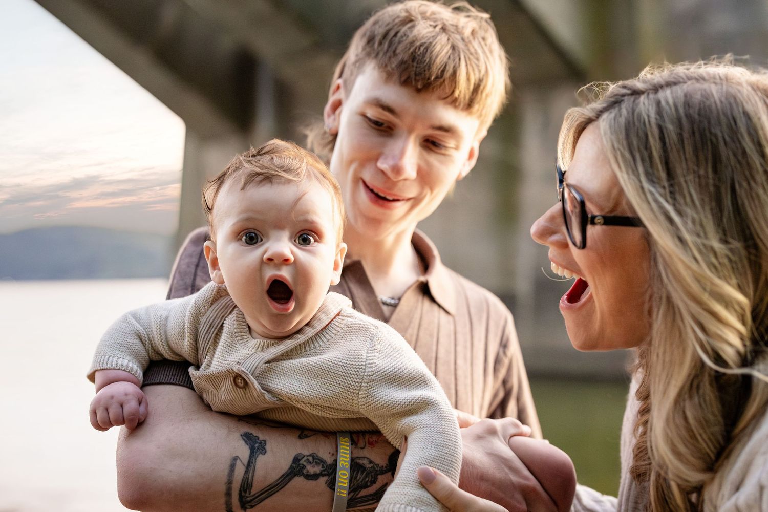 Young couple smiling at wide-eyed baby with anchor tattoo near waterfront bridge.