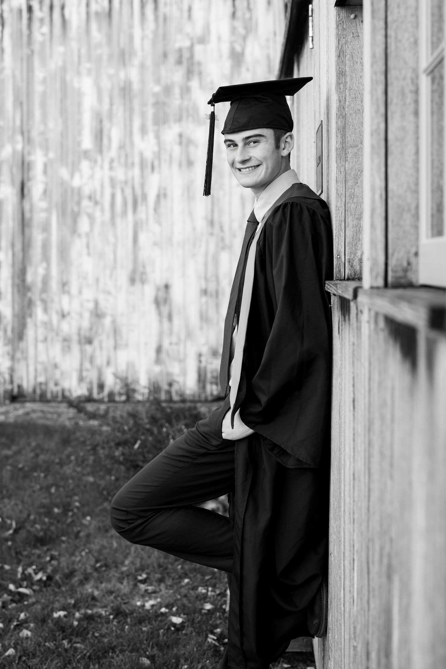 A graduate in traditional black cap and gown leans against a rustic wooden wall in a black and white photo.