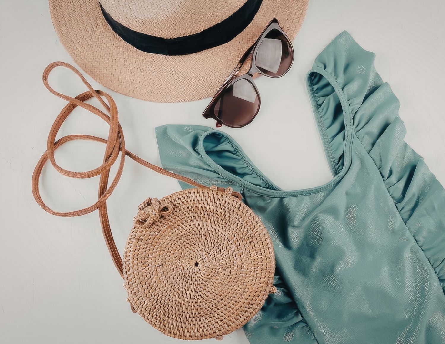 A round woven straw bag with leather strap sits next to a turquoise ruffled garment on white background.