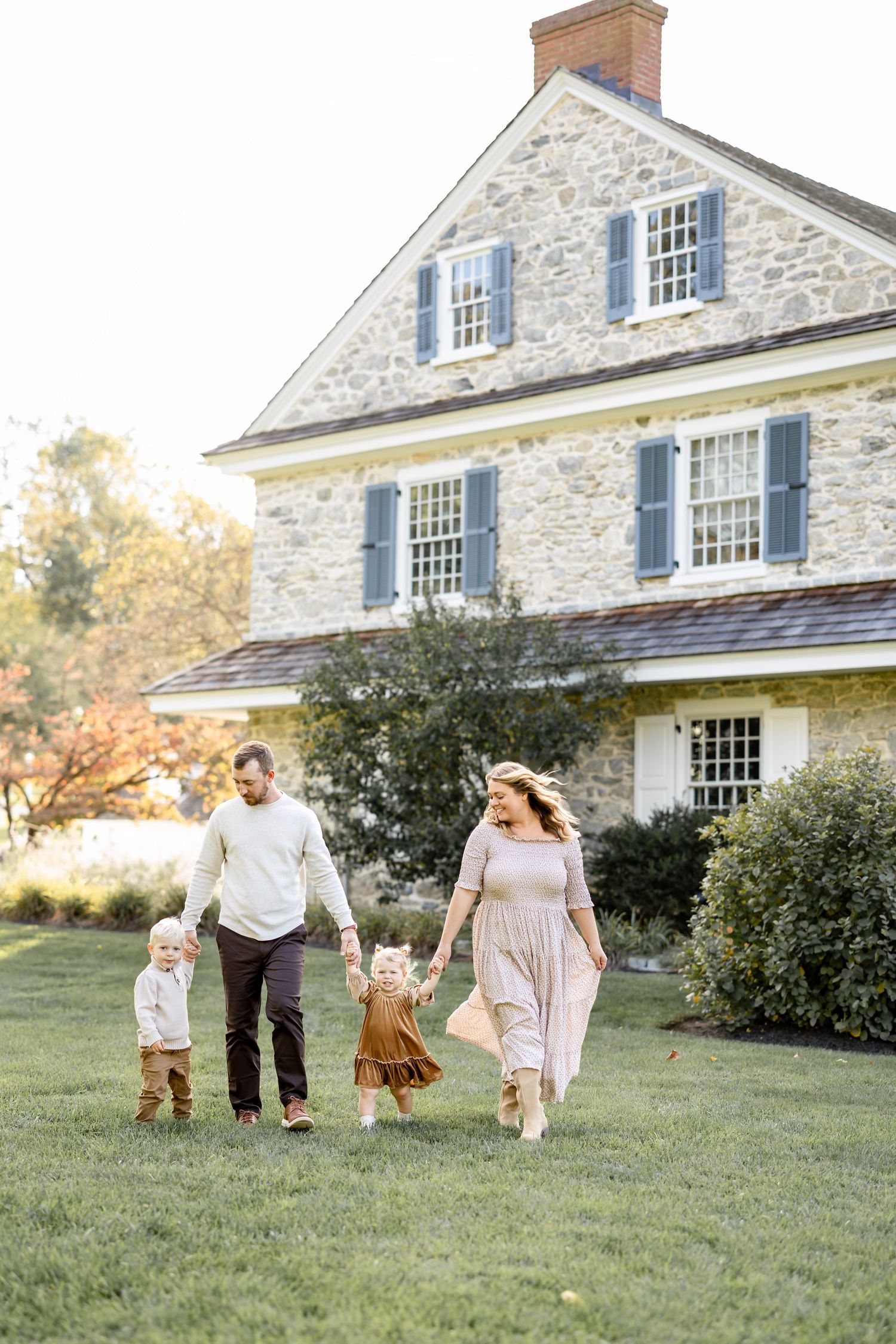 A group dances joyfully on a grassy lawn in front of a historic stone colonial house with white shutters.