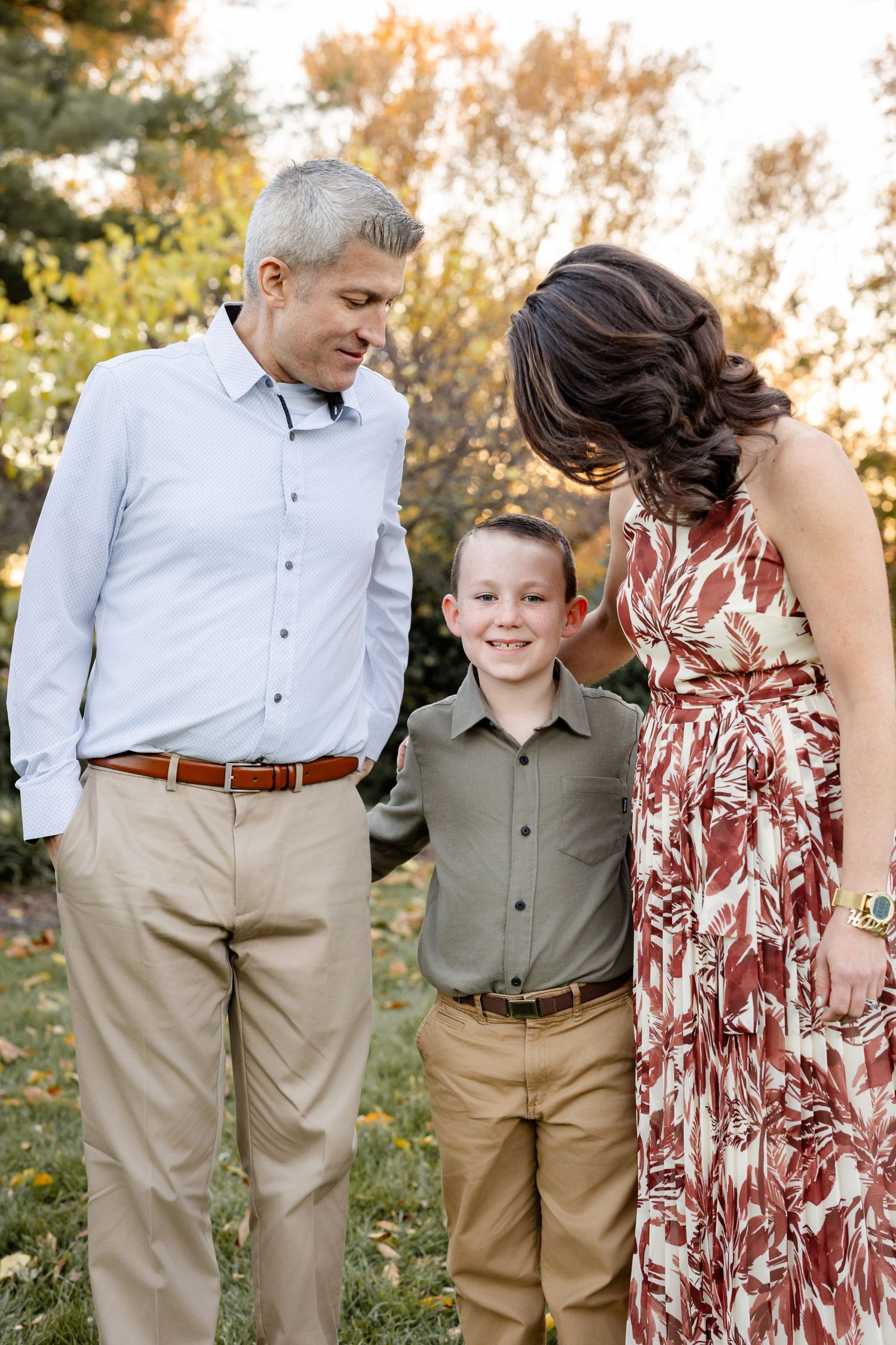 A smiling family of three poses outdoors in autumn, parents looking lovingly at their young son in a park setting.