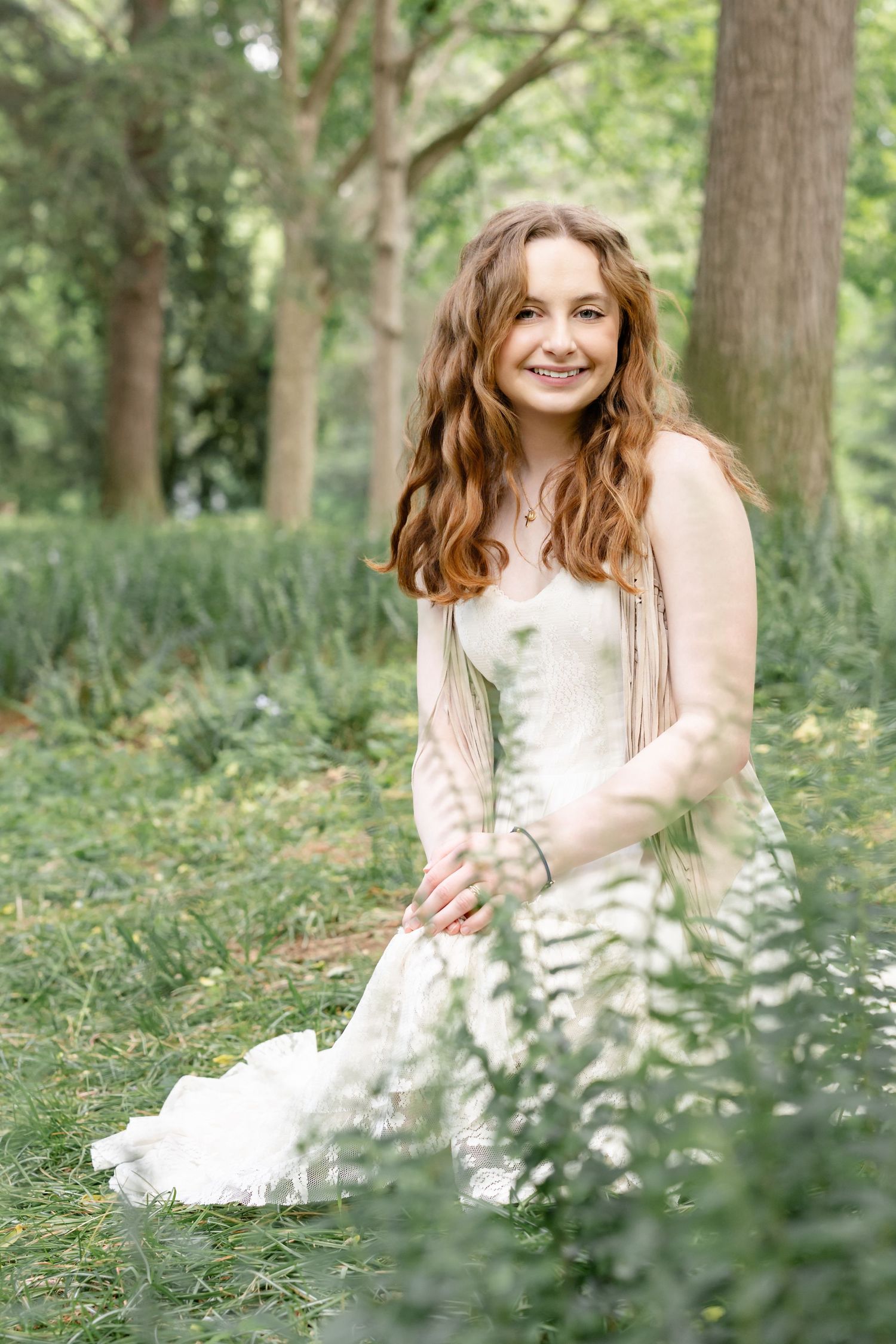 Young woman with long red hair in white dress sitting in lush green park, smiling warmly at camera.