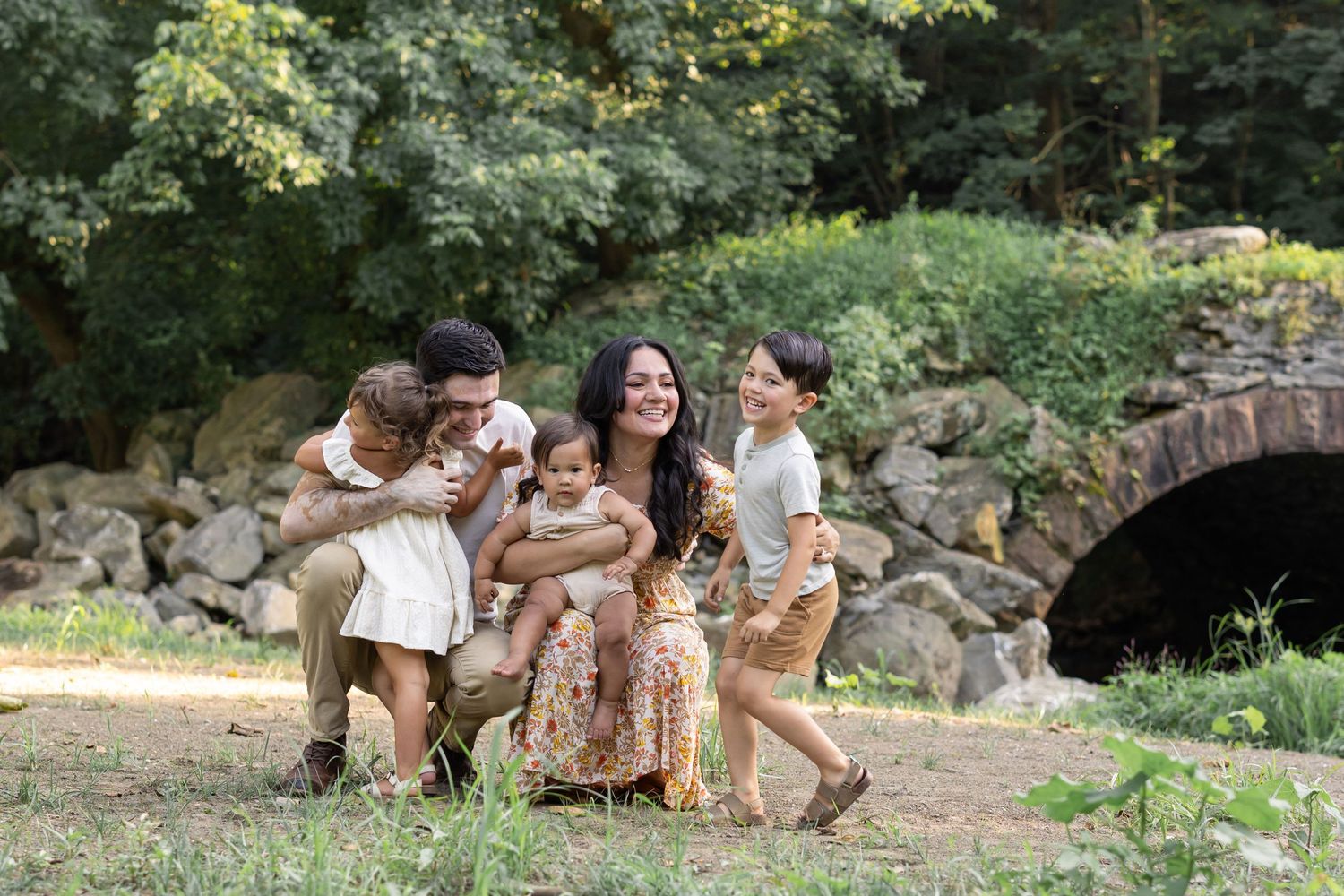 Playful family moment captured in park setting with stone bridge in background.