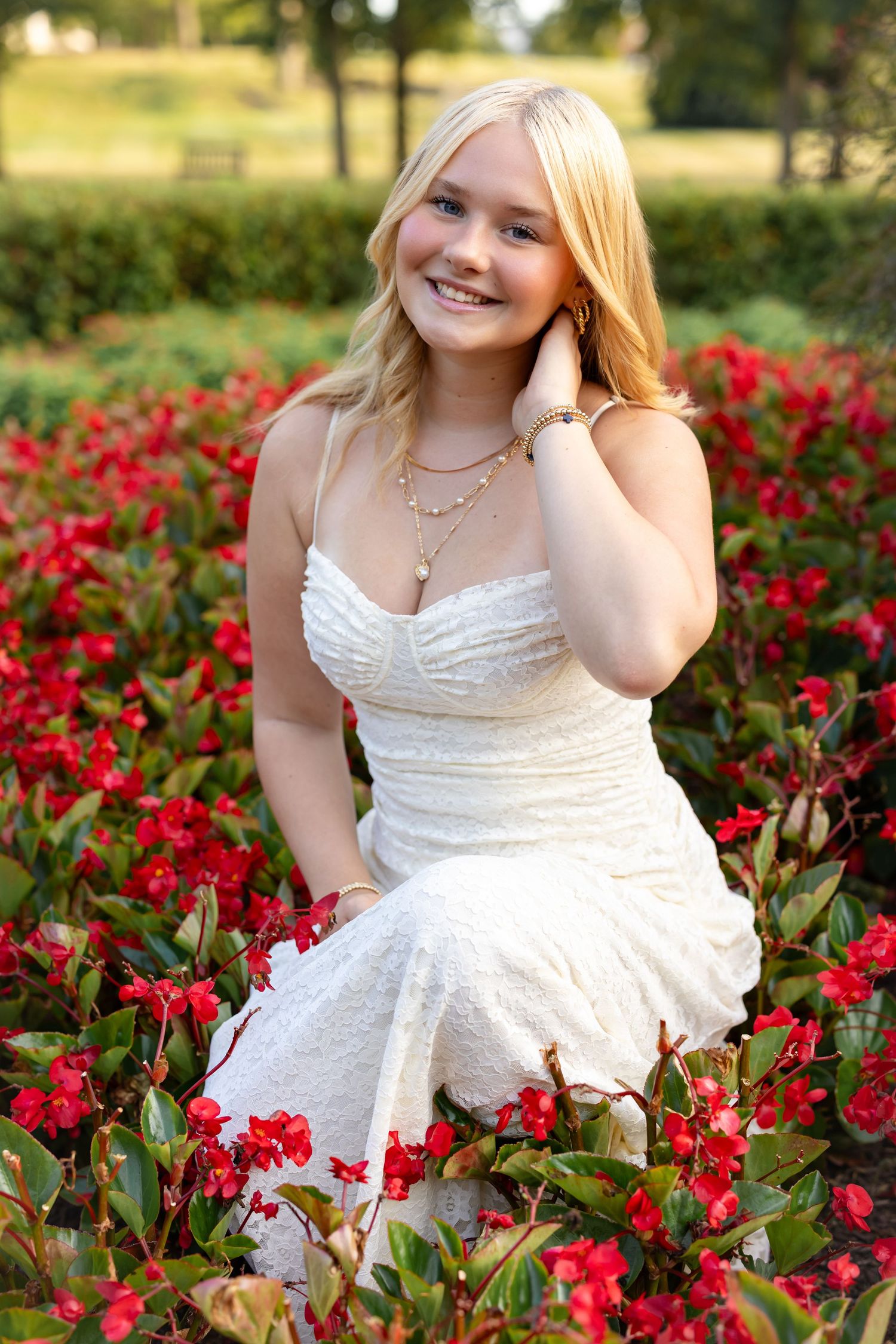 A high school senior wearing a white lace dress stands out beautifully against a backdrop of vibrant red roses in a garden setting.
