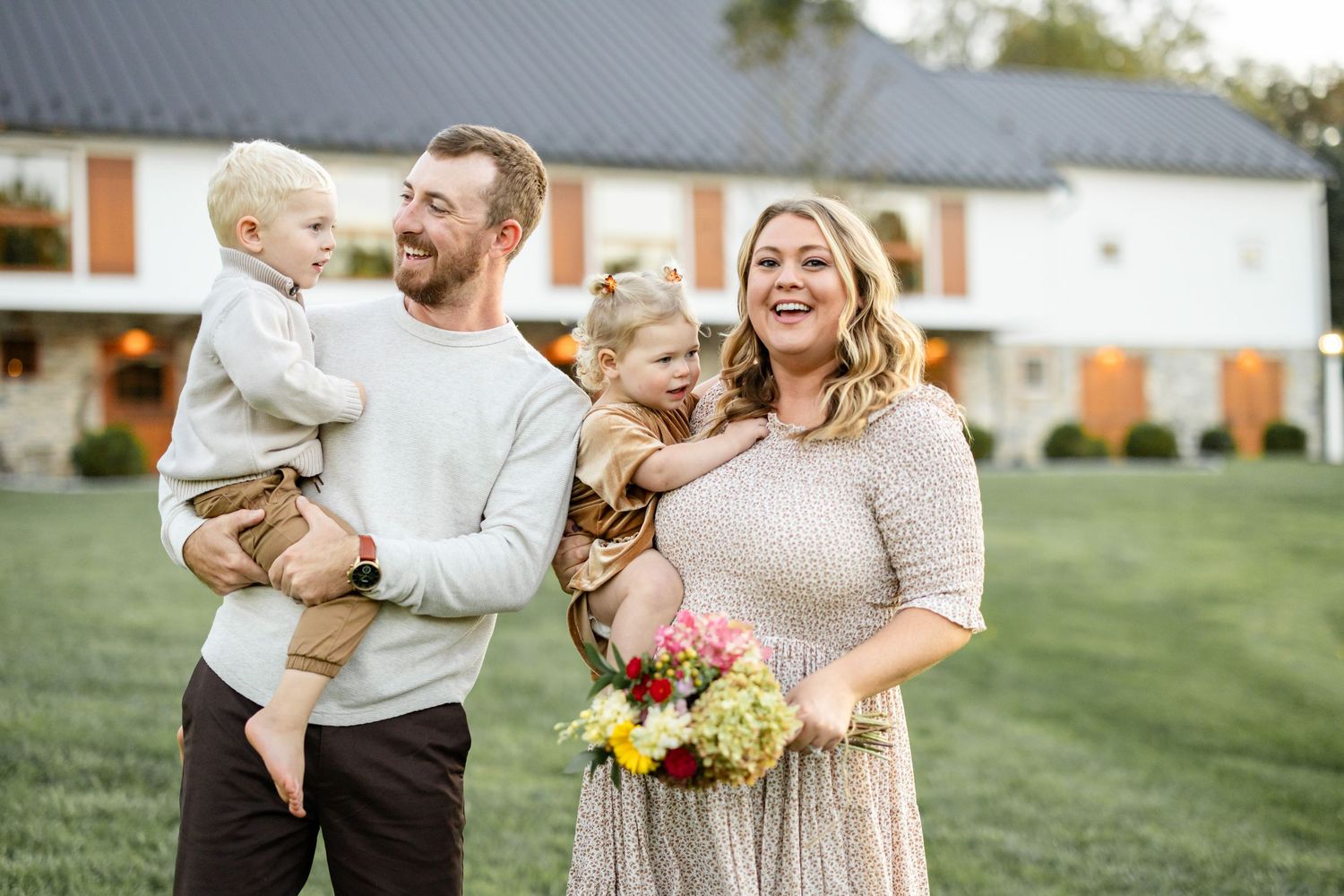 Happy family of four posing outdoors near a white farmhouse, parents holding young children, woman holding colorful flowers.