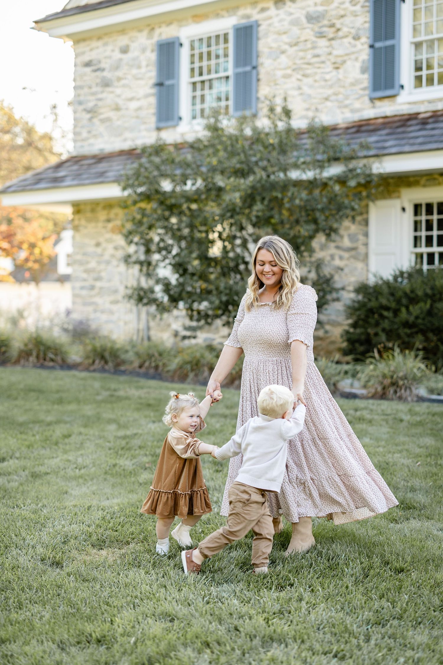 A smiling mother in a flowy dress holds hands with two toddlers on a green lawn outside a stone house.