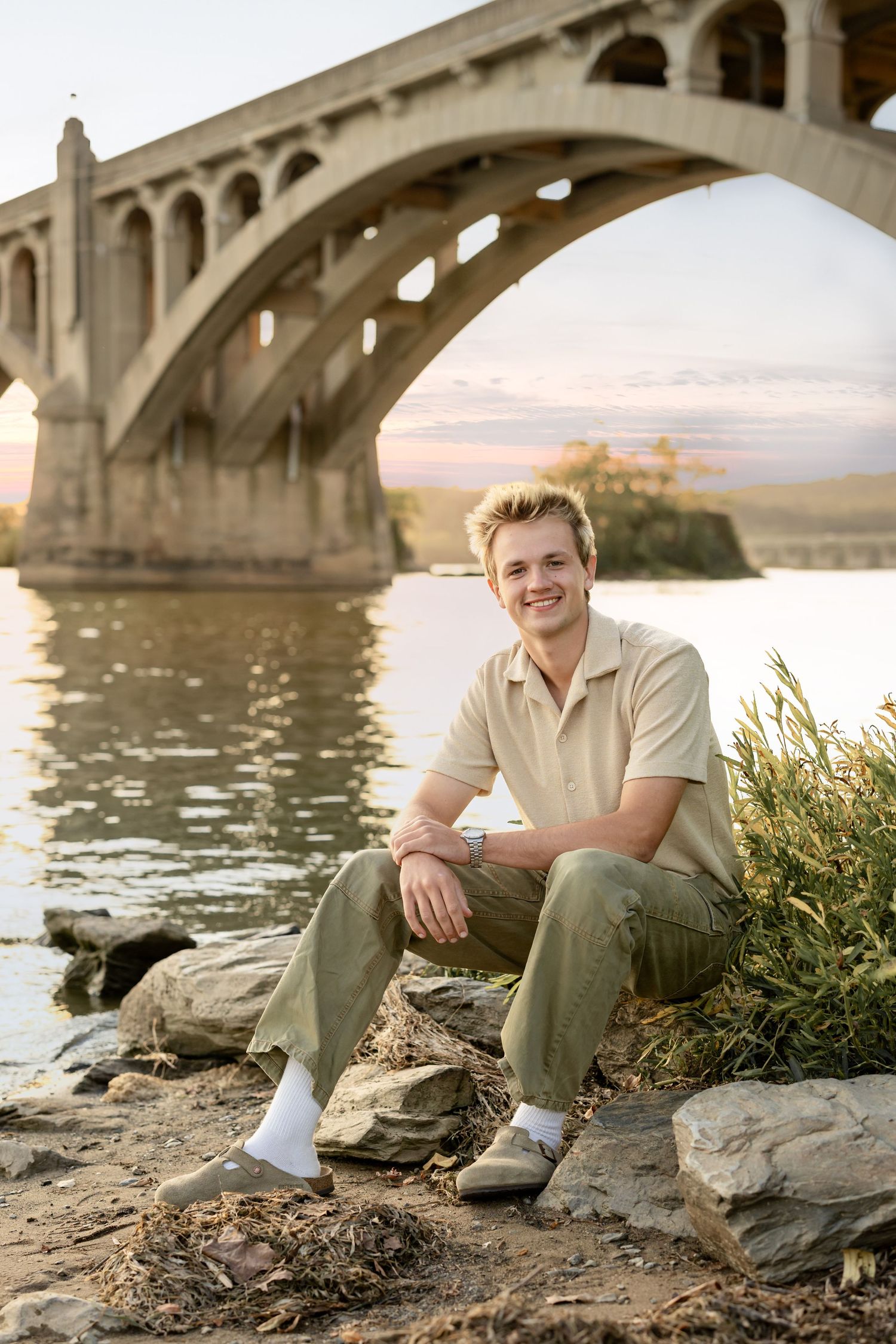 High school senior boy sits casually on rocky riverbank beneath stone bridge during golden sunset hour.