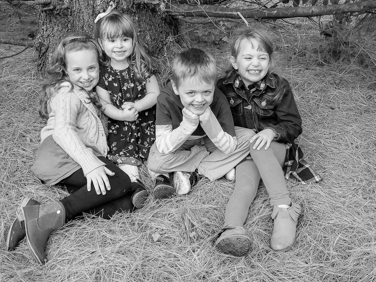 Black and white portrait of four children sitting together outdoors on the ground, smiling and posing playfully.