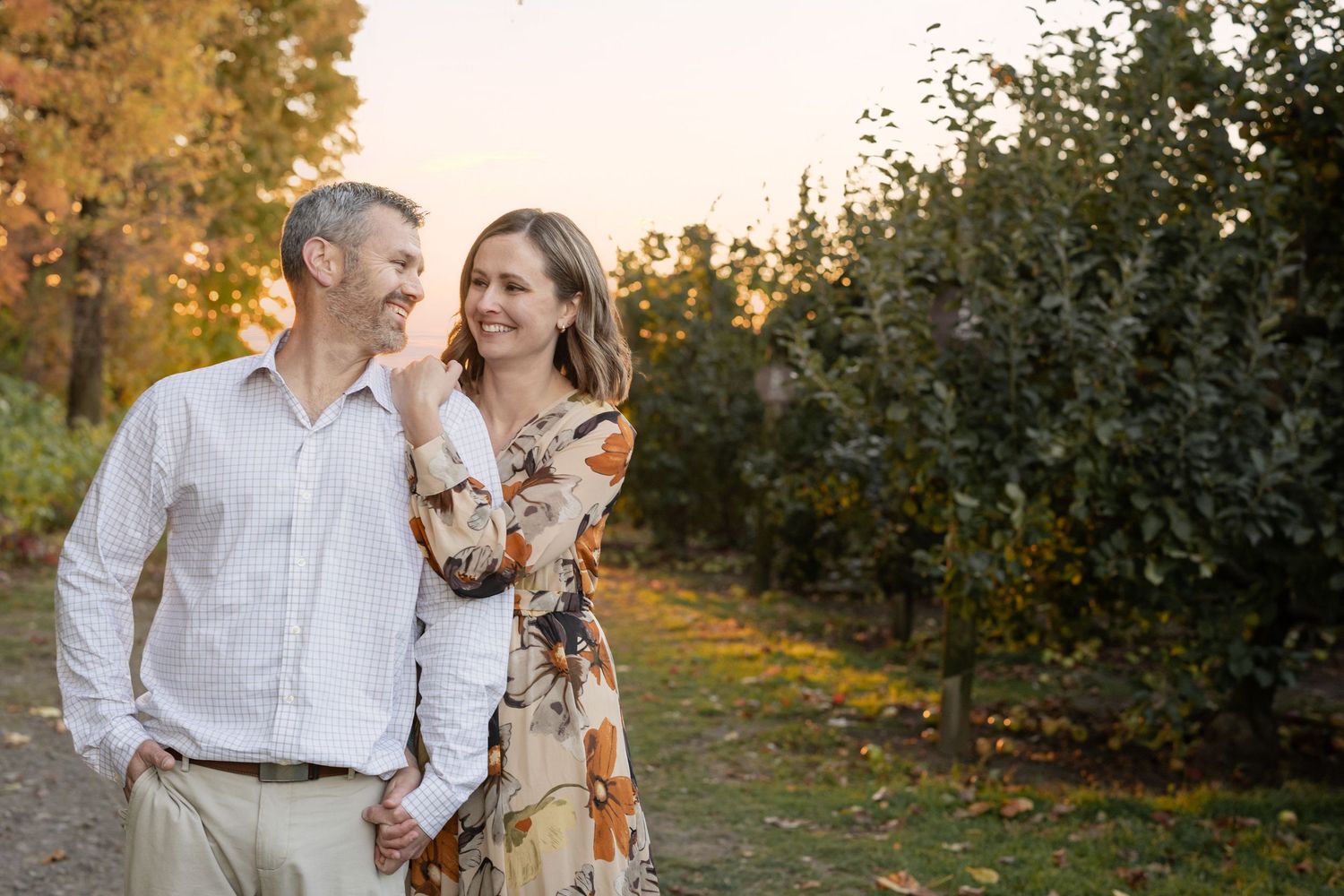 Couple holding hands and smiling in an autumn orchard, wearing elegant attire during golden hour.