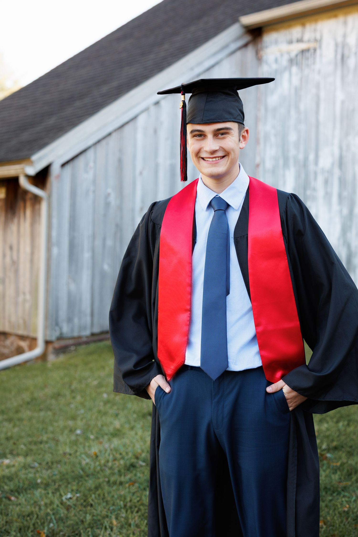 Graduate wearing traditional black cap and gown with red stole stands outdoors in front of rustic barn wall.