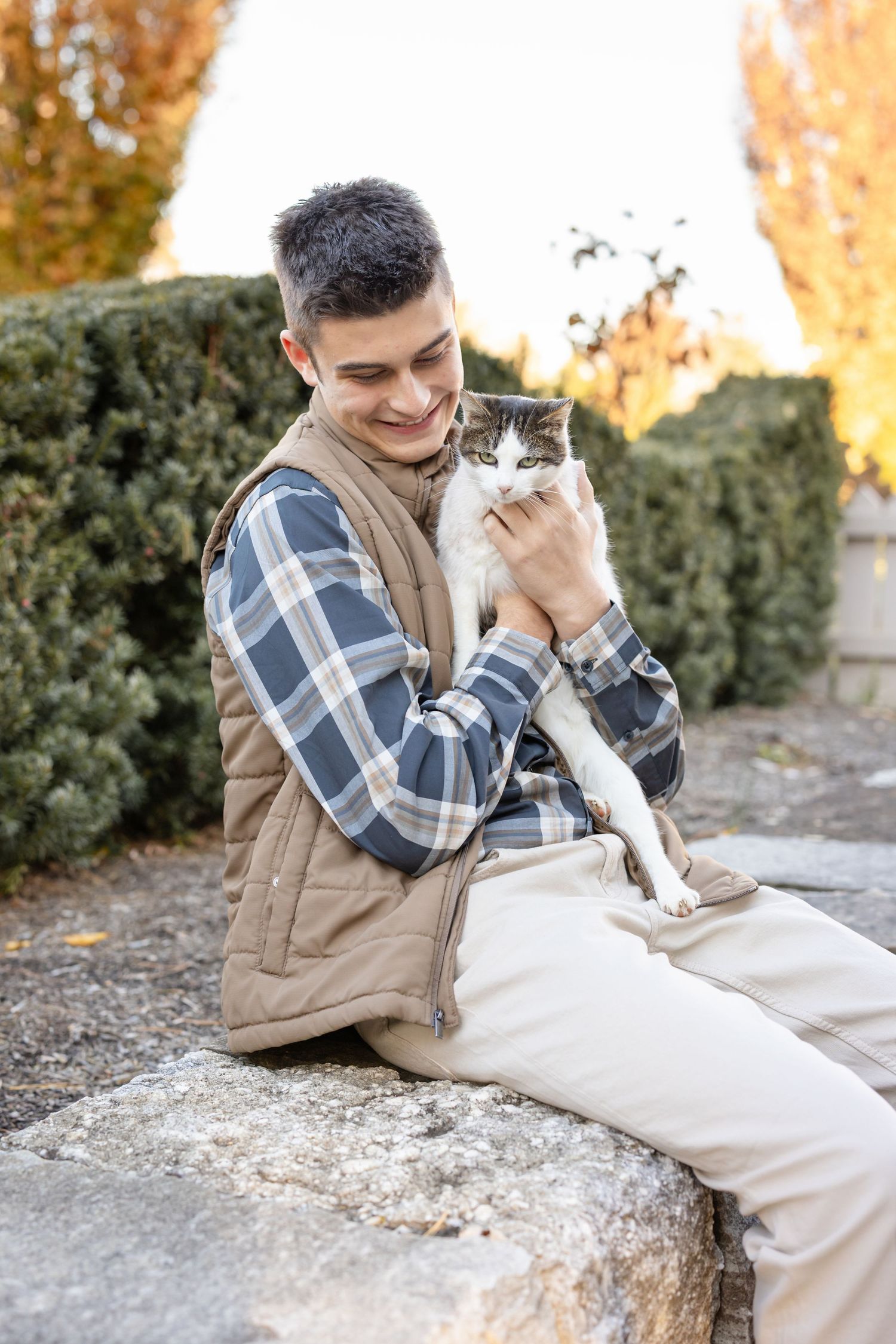 A high school senior guy in a plaid shirt and beige vest cuddles with a white cat on stone steps during a fall afternoon.