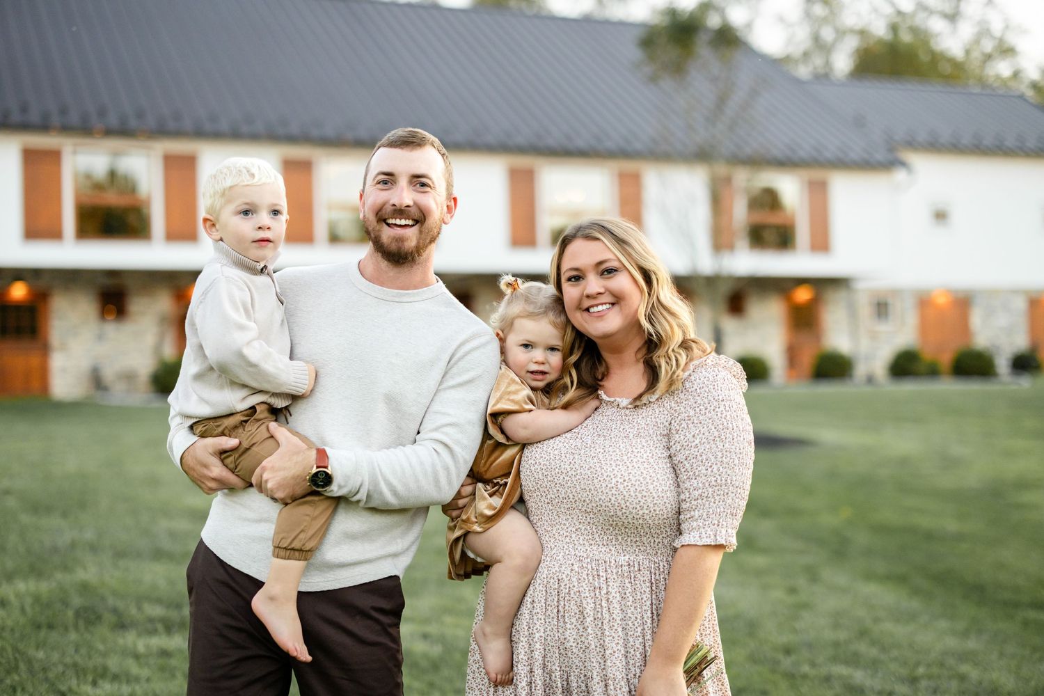 A smiling family of four poses outdoors in front of a white barn, parents holding two young toddlers on a green lawn.