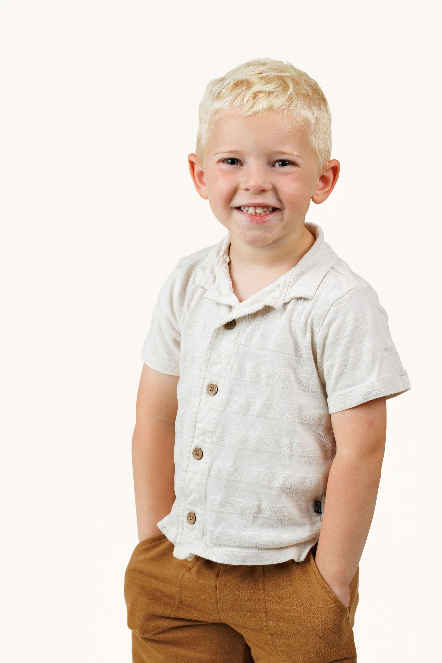 Smiling blonde boy in white button-up shirt and tan pants posing with hands in pockets against white background.