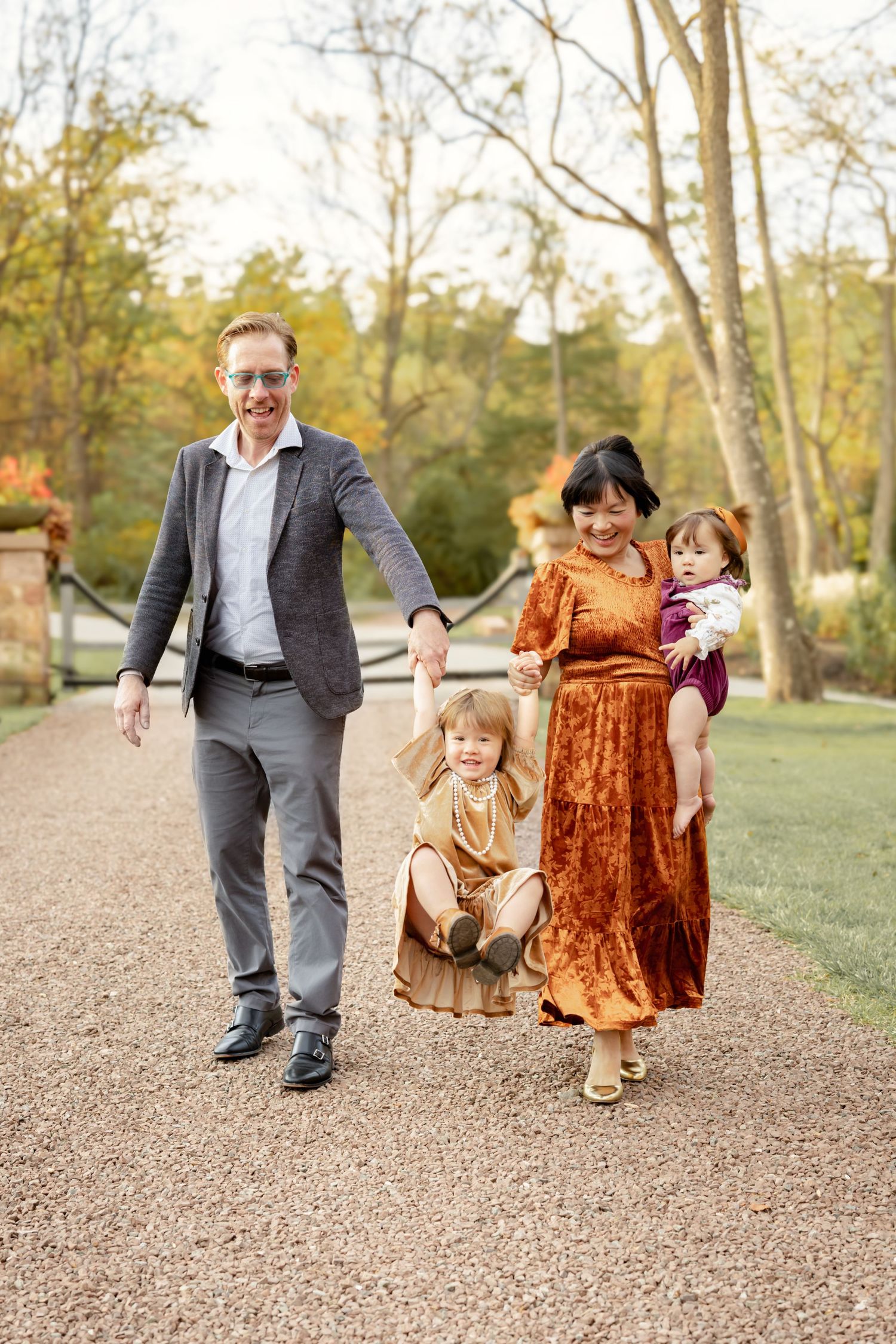 A joyful family of four walks along a gravel path in an autumn park, dressed in elegant fall-toned outfits.