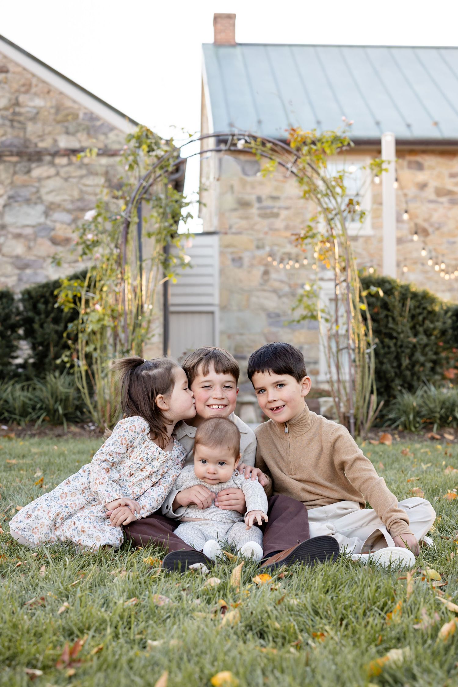 Four young siblings sit together on green grass in front of a stone building with a metal garden arch.
