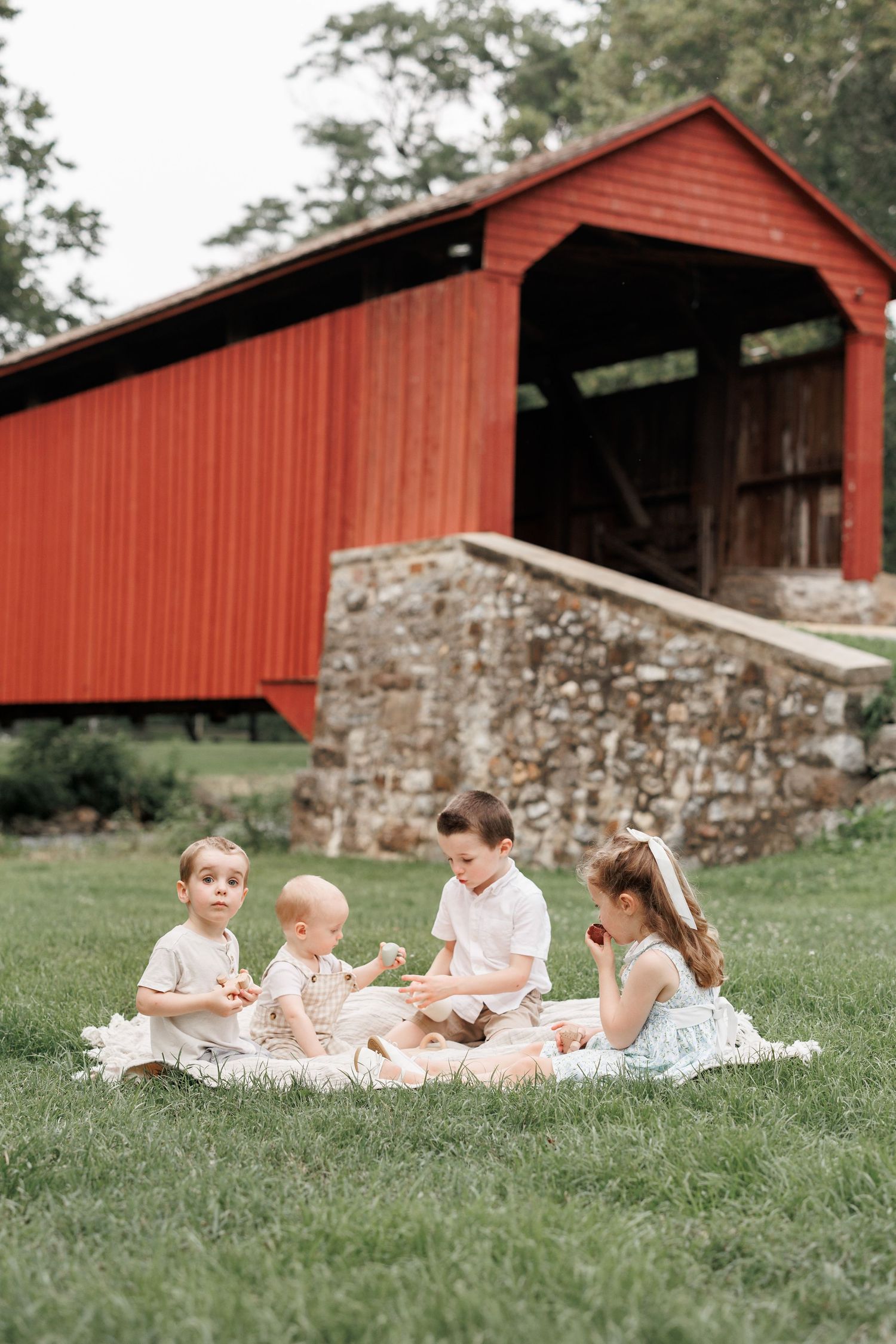 Four young children sit on a blanket in front of a red covered bridge on a lush green lawn.