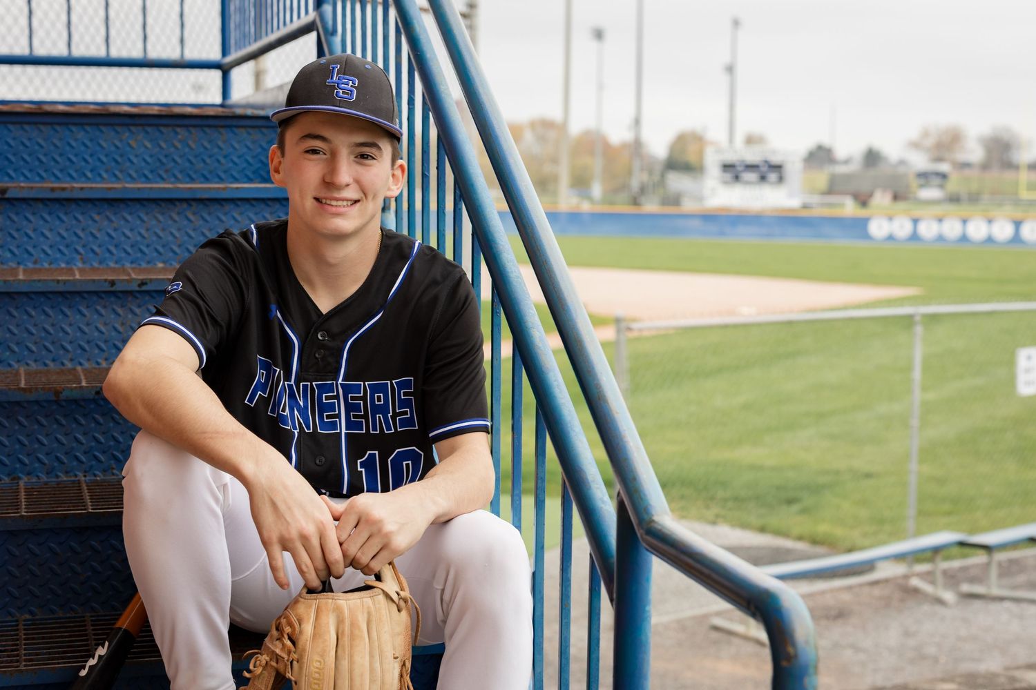 Baseball player sits on dugout steps wearing a navy cap and black uniform during a game at an outdoor field.