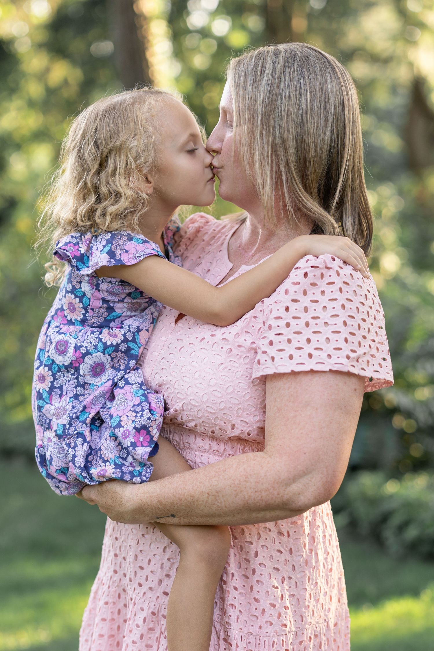 A mother and daughter in pink and purple dresses share a sweet kiss in a beautiful garden, image by Lancaster, PA family photographer.