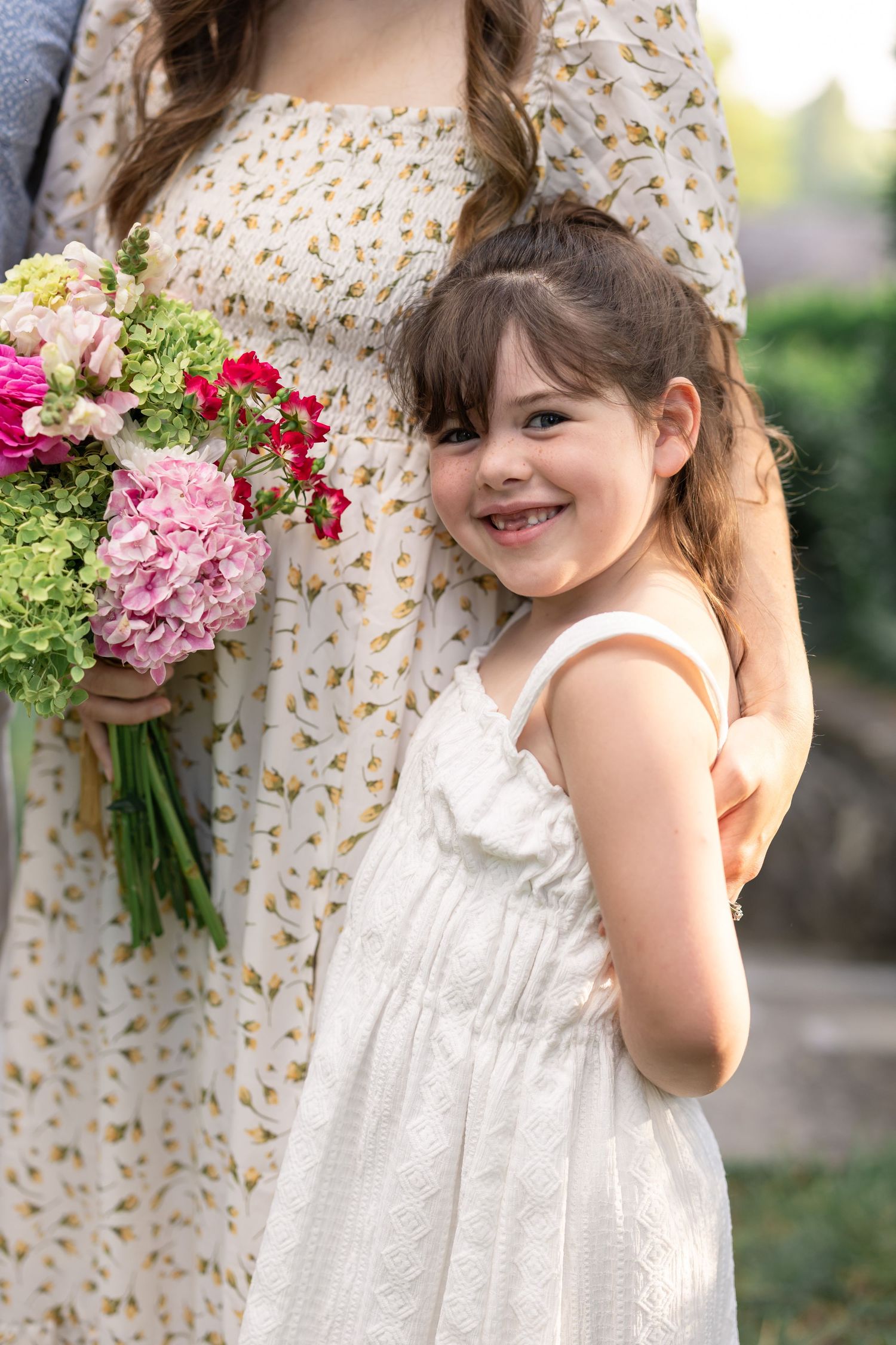 A smiling little girl in a white dress stands beside a woman holding a pink and green floral bouquet outdoors.