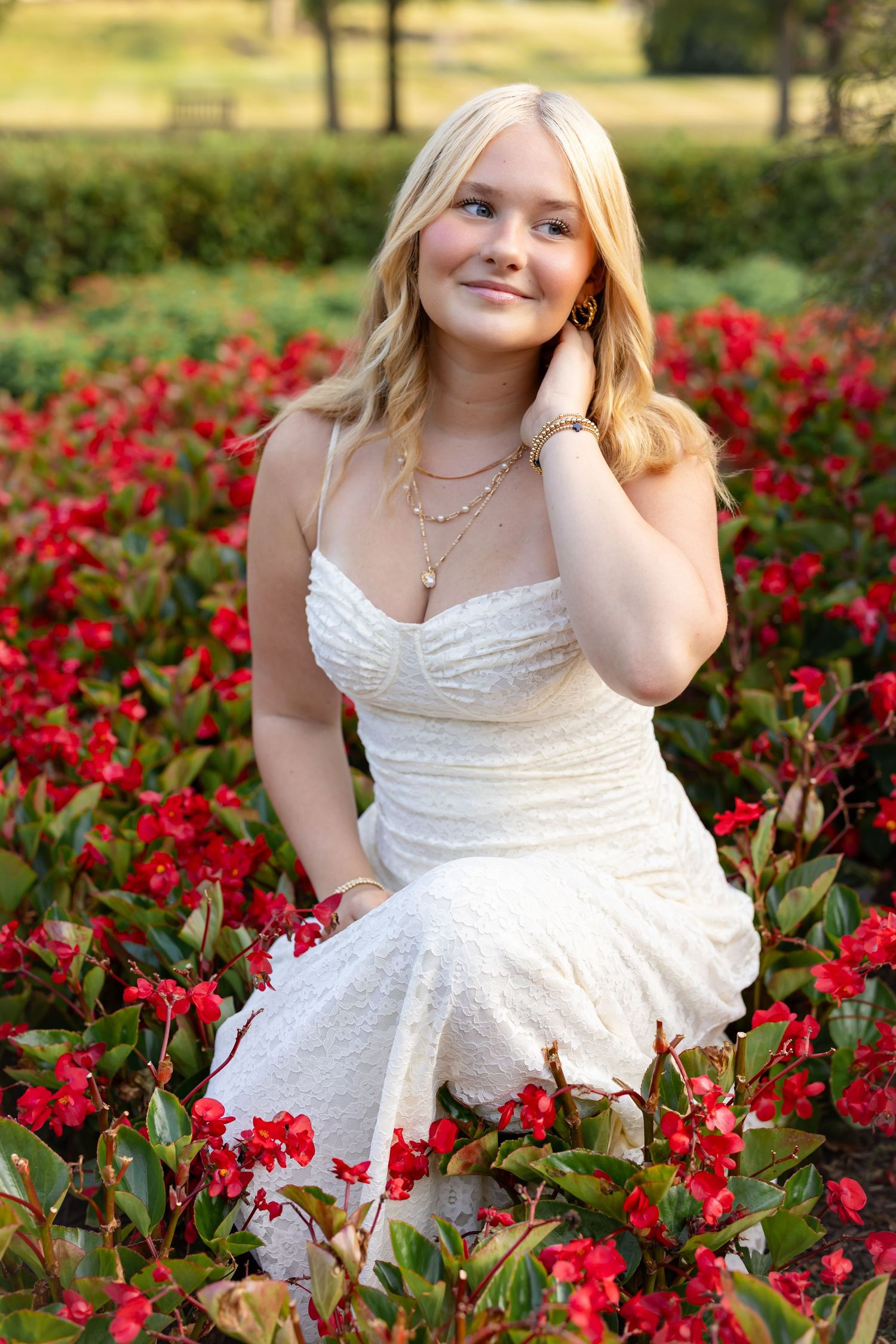 A high school senior girl n in a white dress poses gracefully among vibrant red roses in a garden setting.