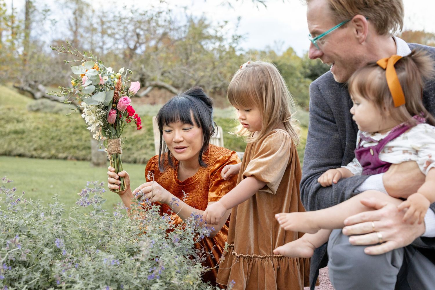 Woman and girl arranging a floral bouquet outdoors while a man holds a baby in a lush garden setting.