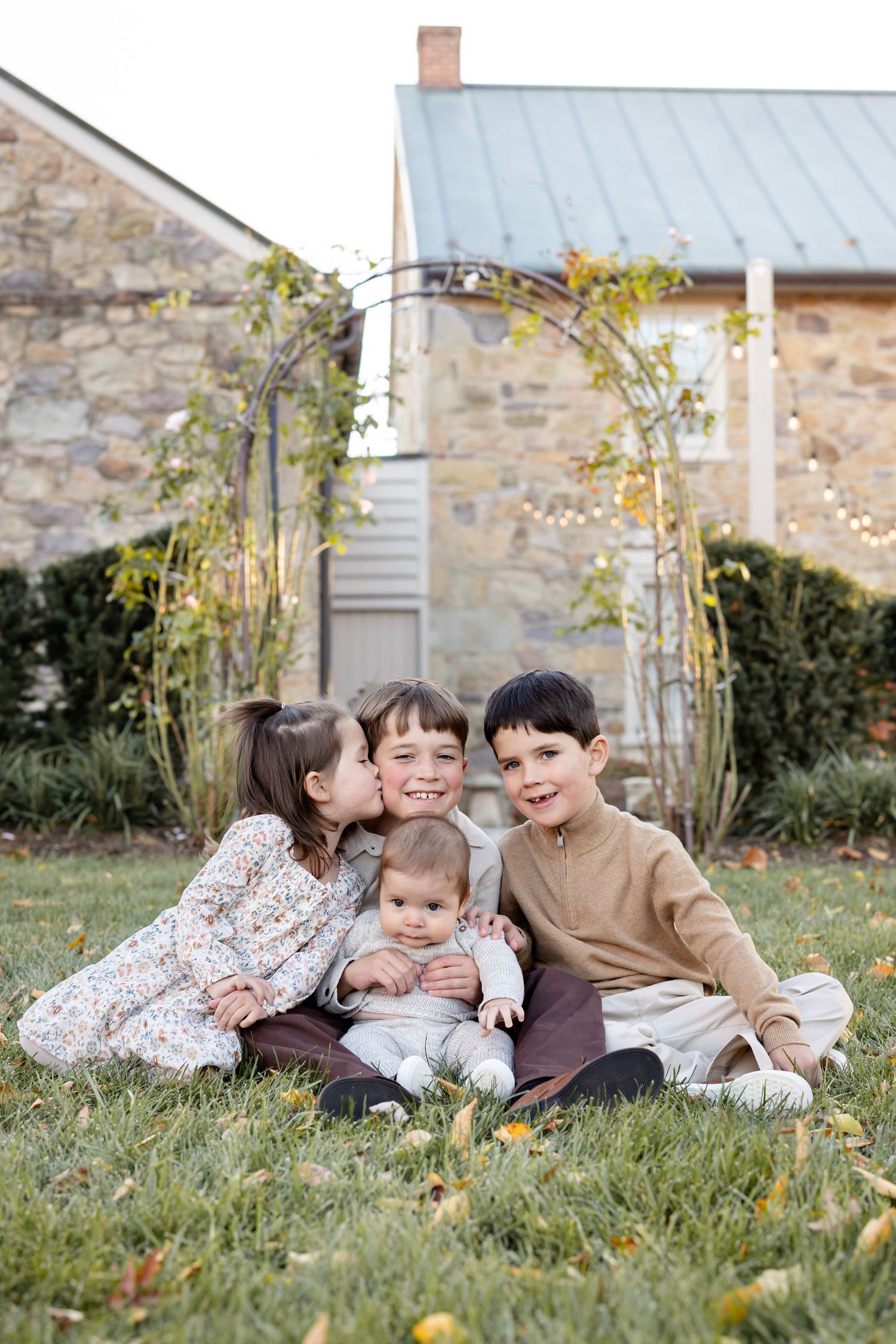 Four siblings sit together on grass outdoors, with older kids hugging a baby, in front of a stone building with a garden arch.