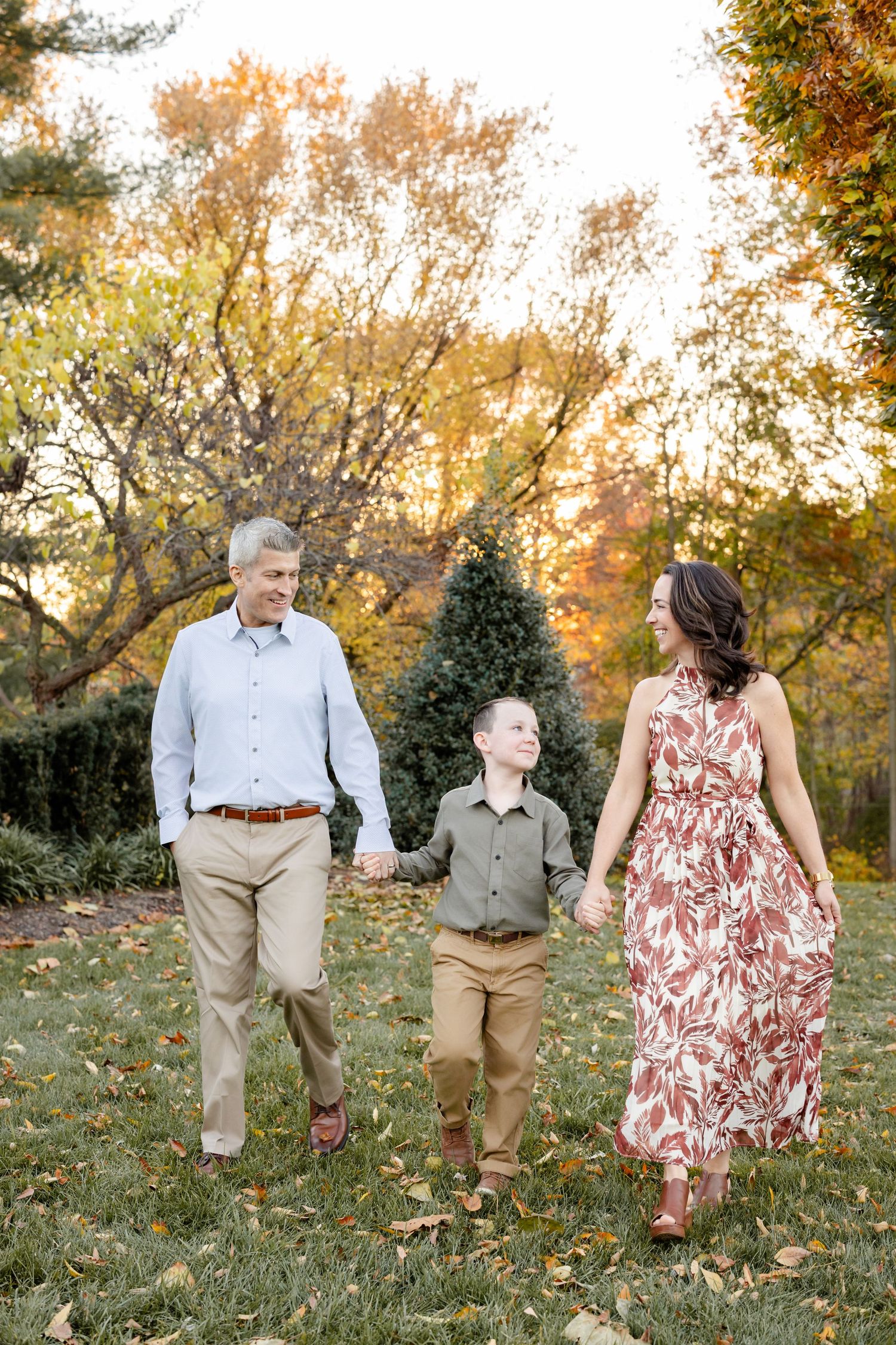 Three people walk together through fallen autumn leaves in a park setting during golden hour.