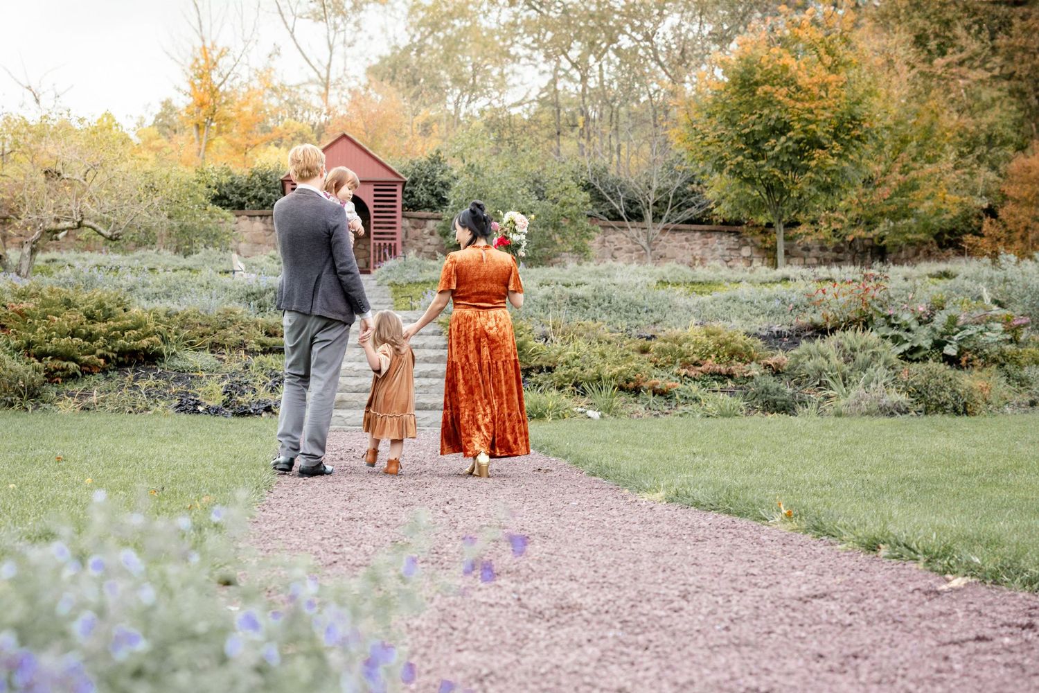 People in autumn attire walk along a winding path through a scenic garden with fall foliage and rustic buildings.
