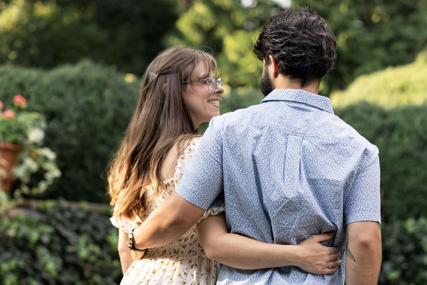 A couple embraces outdoors in a garden, smiling at each other, woman wearing glasses and floral dress.