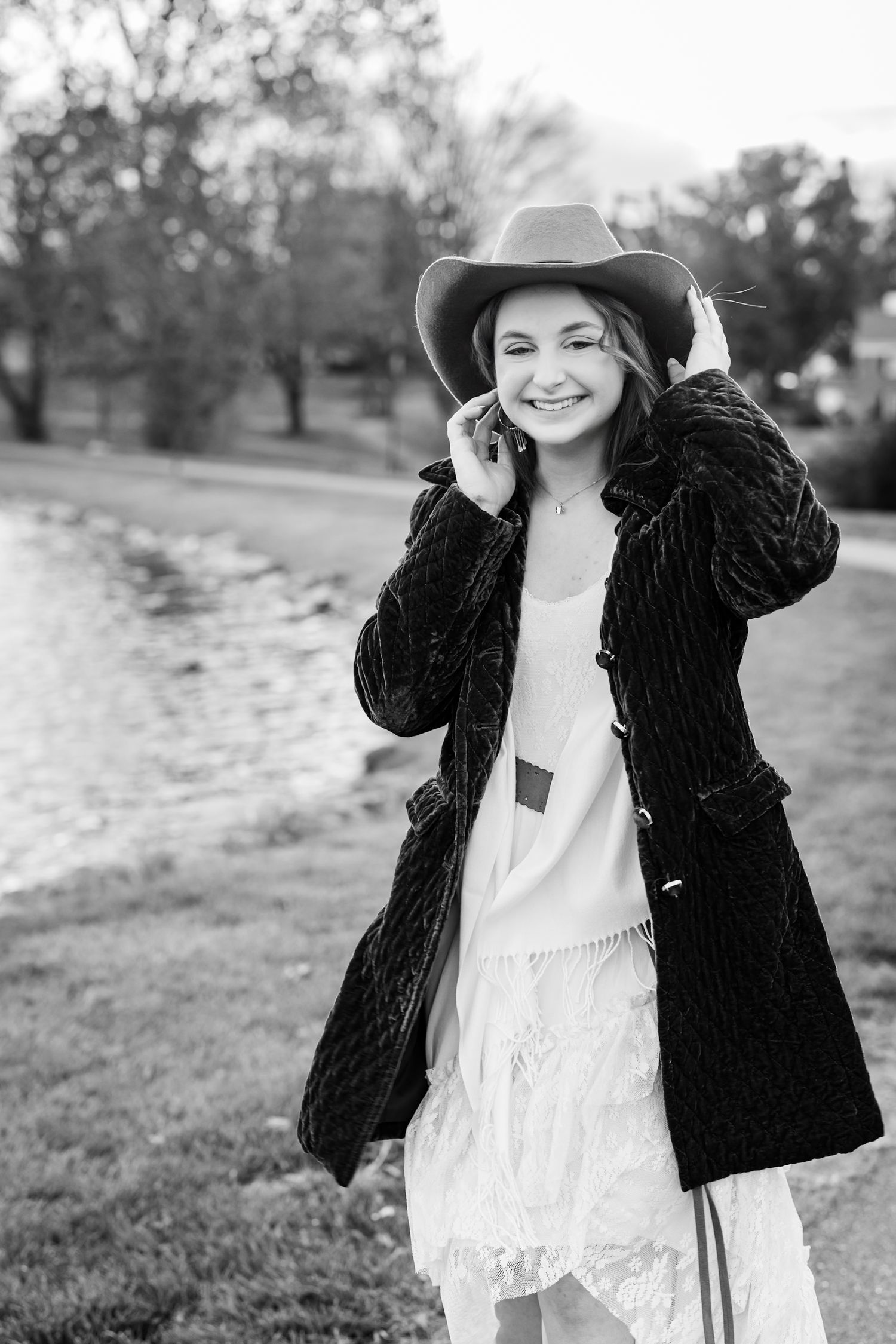 Senior girl smiling in dark jacket and white dress posing outdoors in a natural setting for senior pictures.