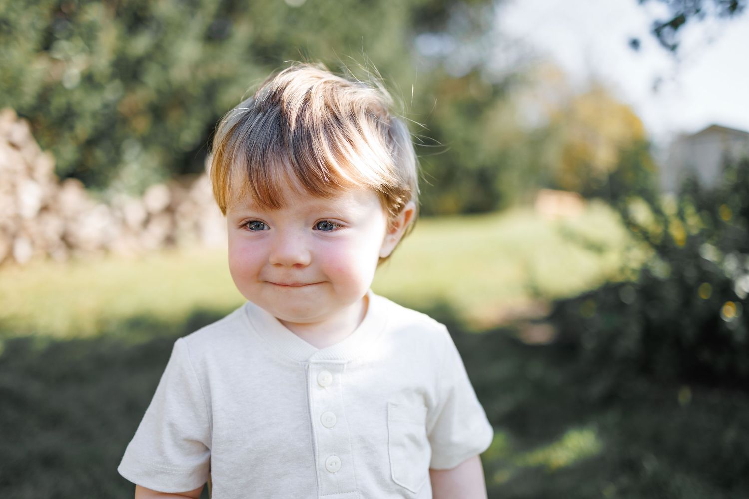 Smiling toddler boy with light hair wearing a white shirt, standing outdoors in a sunny green garden.