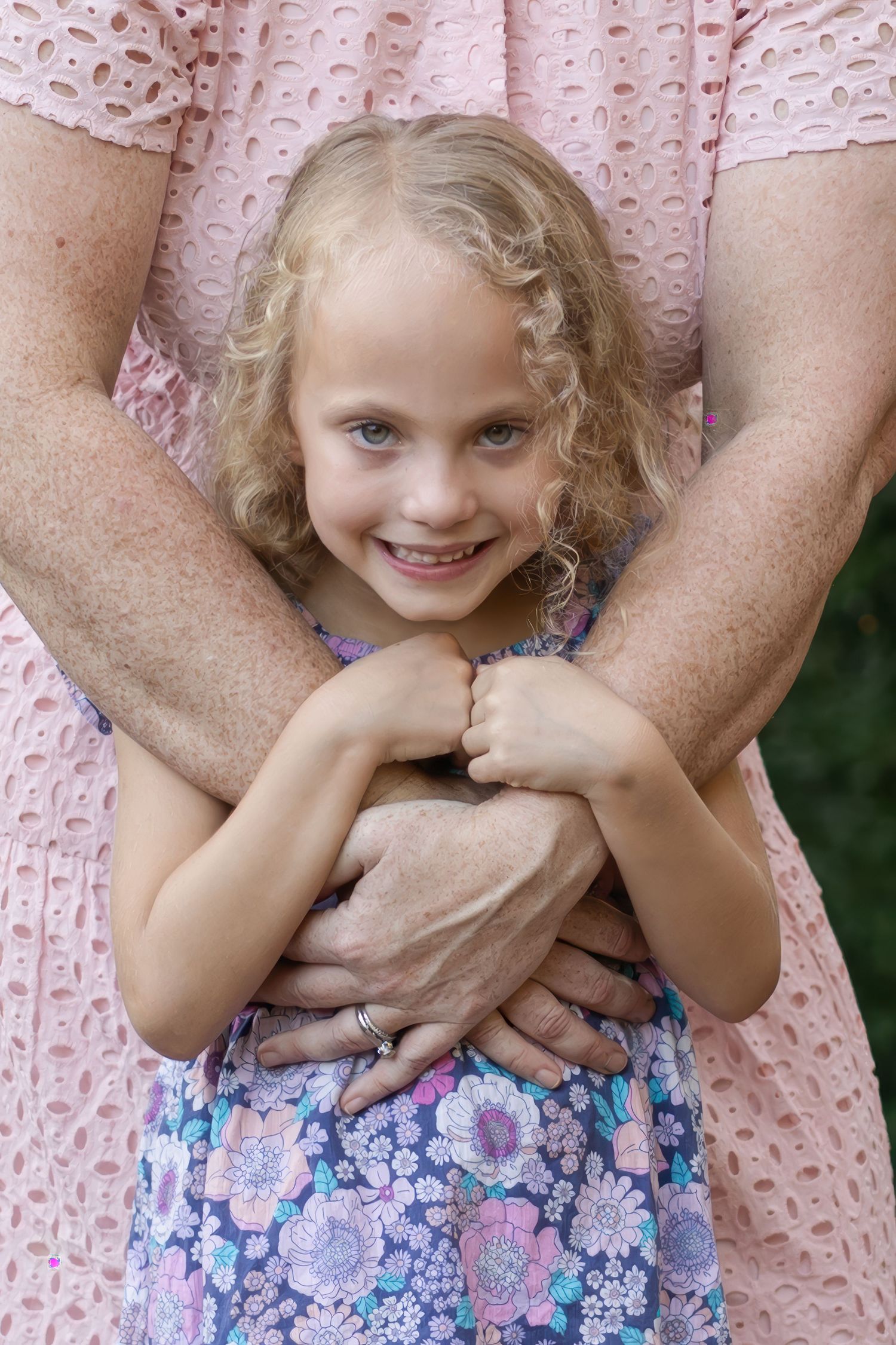 Young blonde girl in floral dress smiling while being hugged from behind by an adult in a pink lace dress.