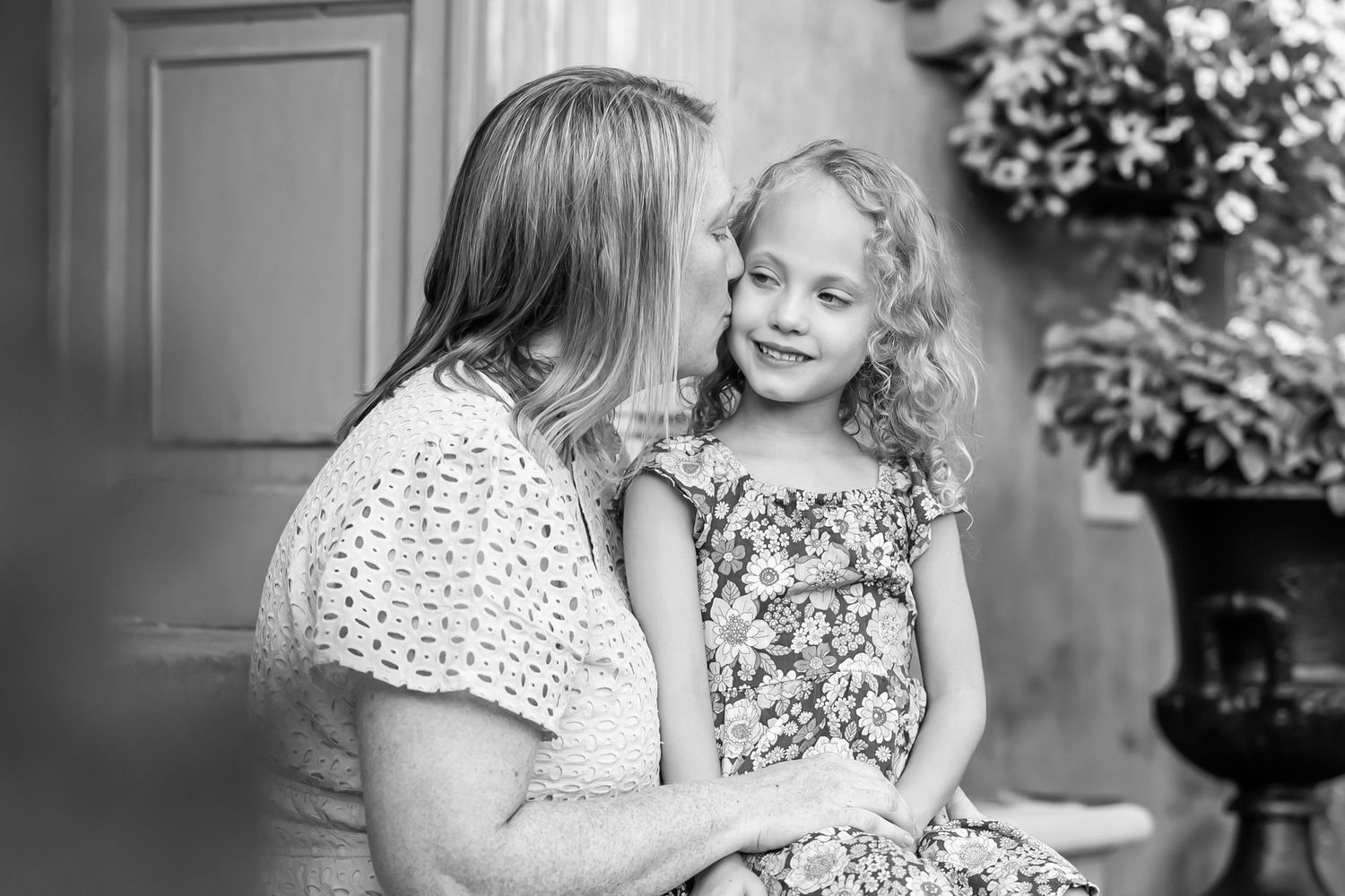 Black and white photo of a mother whispering to her smiling young curly-haired daughter outdoors.