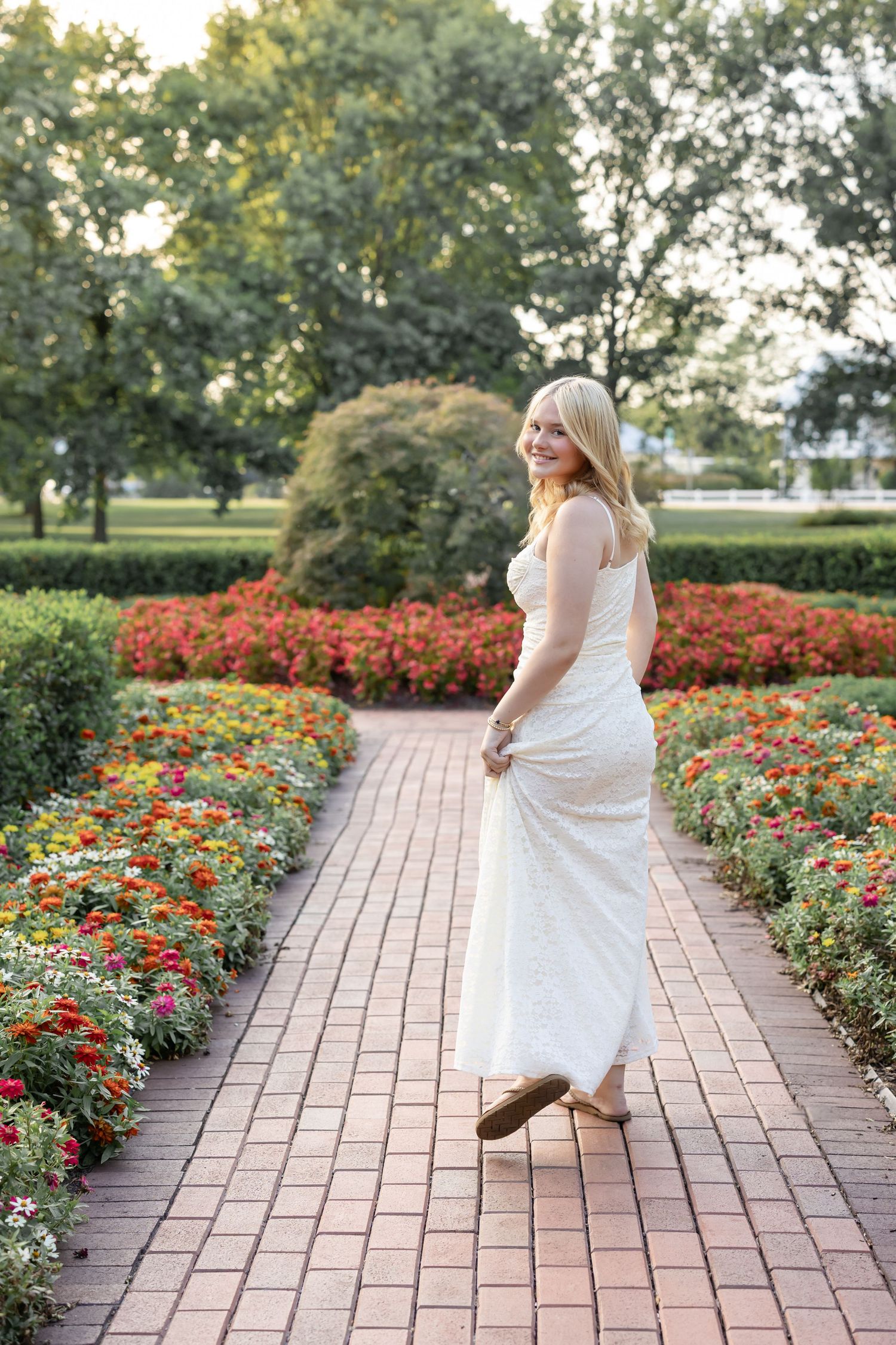 High school senior girl in white dress twirls joyfully on a brick pathway surrounded by vibrant red and orange flower gardens.