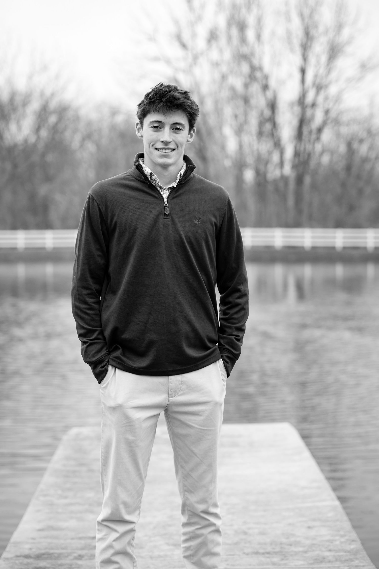 Black and white portrait of a high school boy standing on a wooden dock wearing a dark pullover and light pants near a lake.