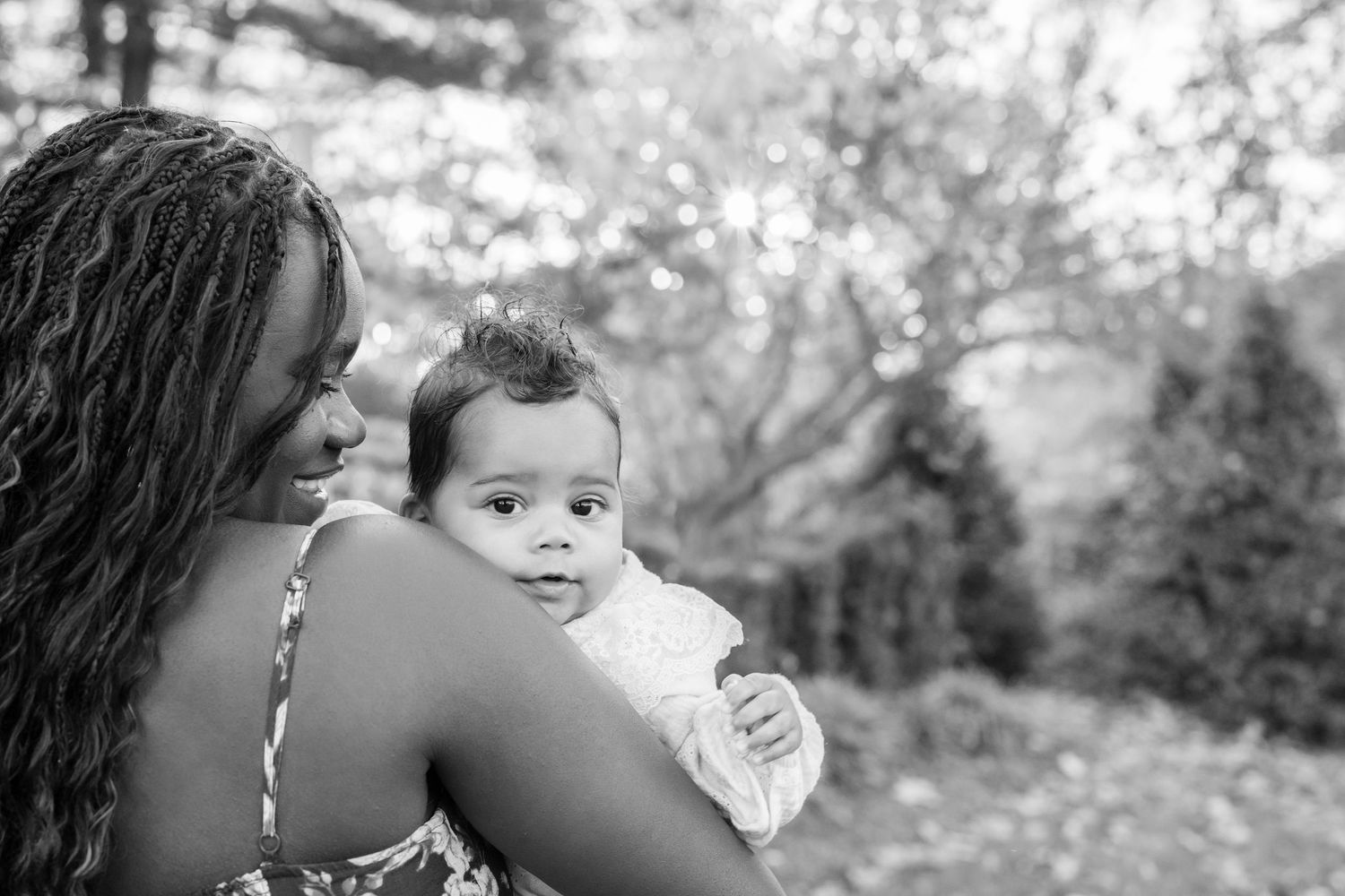 Black and white photo of a mother tenderly holding her baby outdoors, with blurred trees in the background.