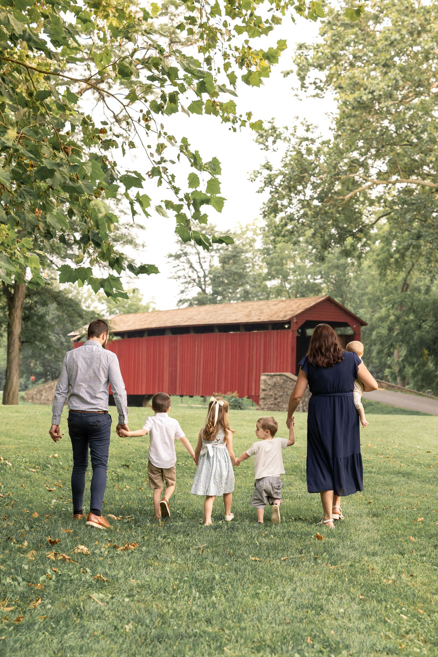 A family walks hand in hand through a grassy field with a red barn in the background on a sunny summer day.
