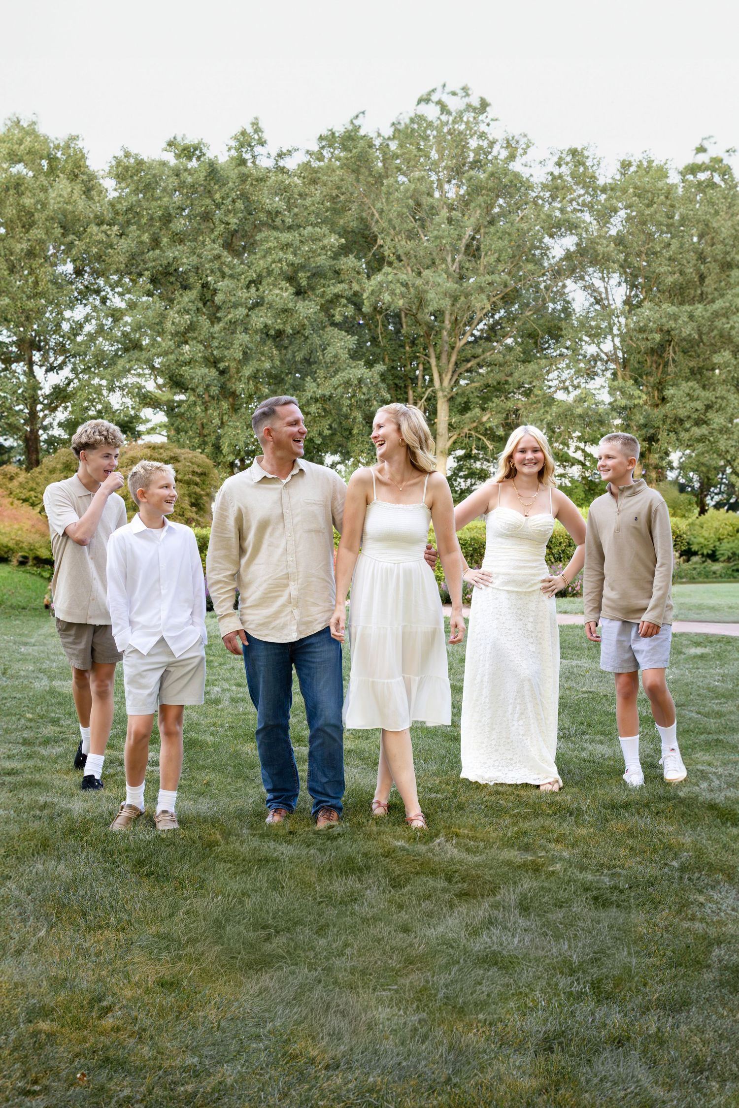 Happy family of six walking together on green grass, wearing neutral tones, smiling outdoors with trees in background.