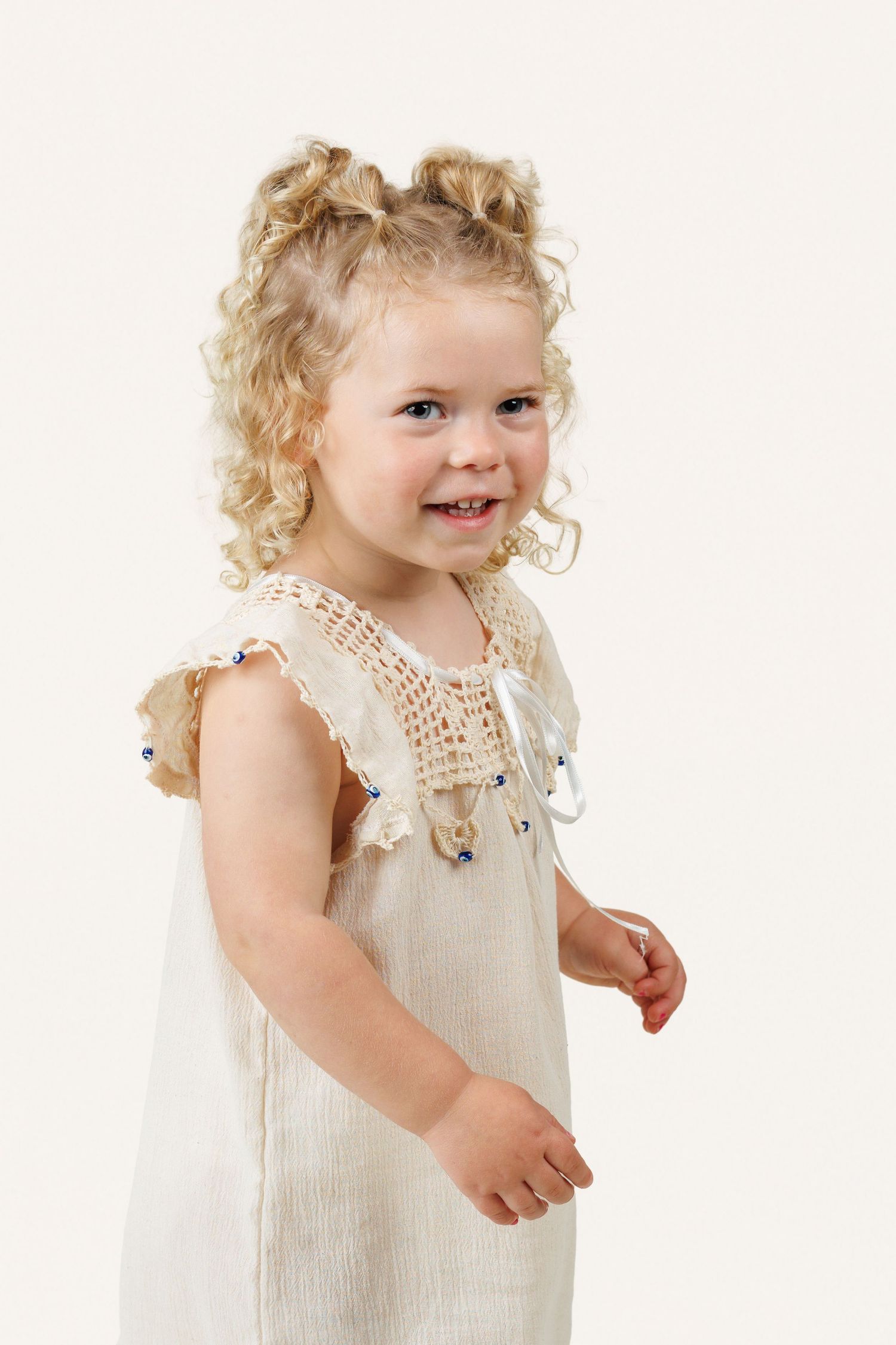 Smiling toddler girl with curly blonde hair in a cream linen dress with crochet collar, posing on white background.